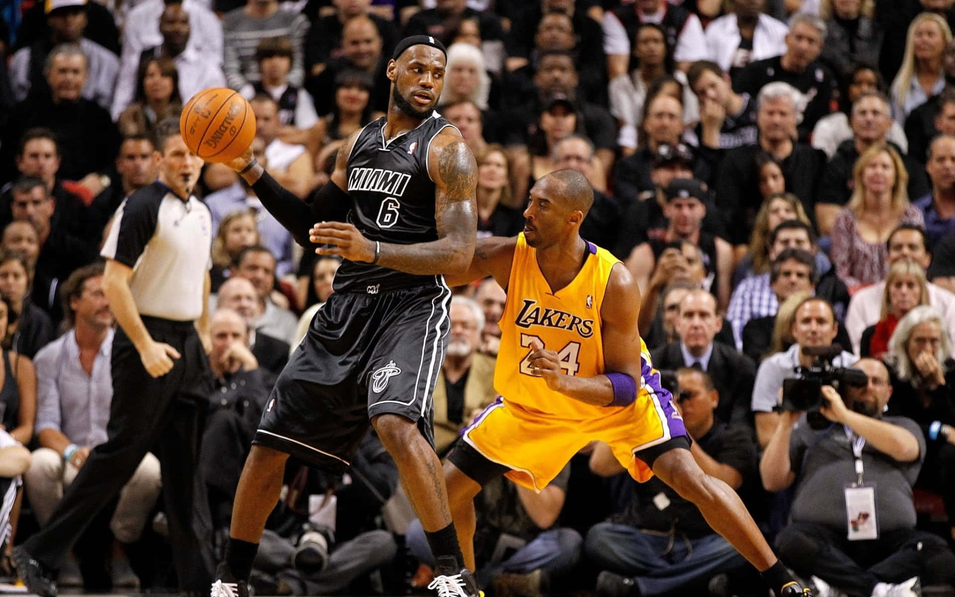NBA巨星聯手閃耀！ 壁紙 — 勒邦占士身穿邁阿密熱火球衣運球，同時被身穿湖人球衣的高比拜仁緊密防守，在一場座無虛席的籃球比賽中。