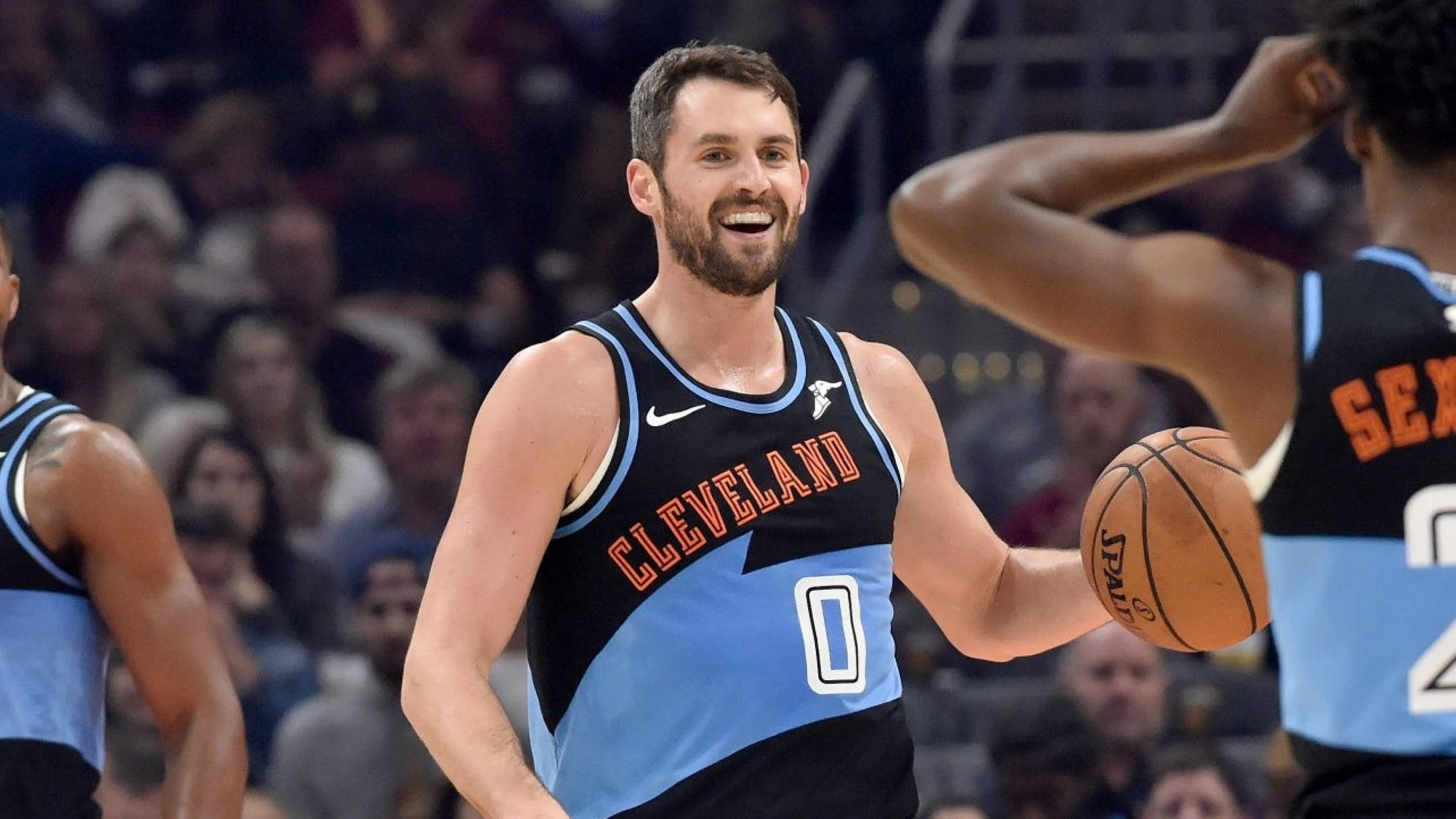 NBA球員 凱文·洛夫 壁紙 — 一位微笑的克里夫蘭籃球運動員身穿藍黑球衣，在激烈的比賽中手持籃球，背景有隊友和觀眾。