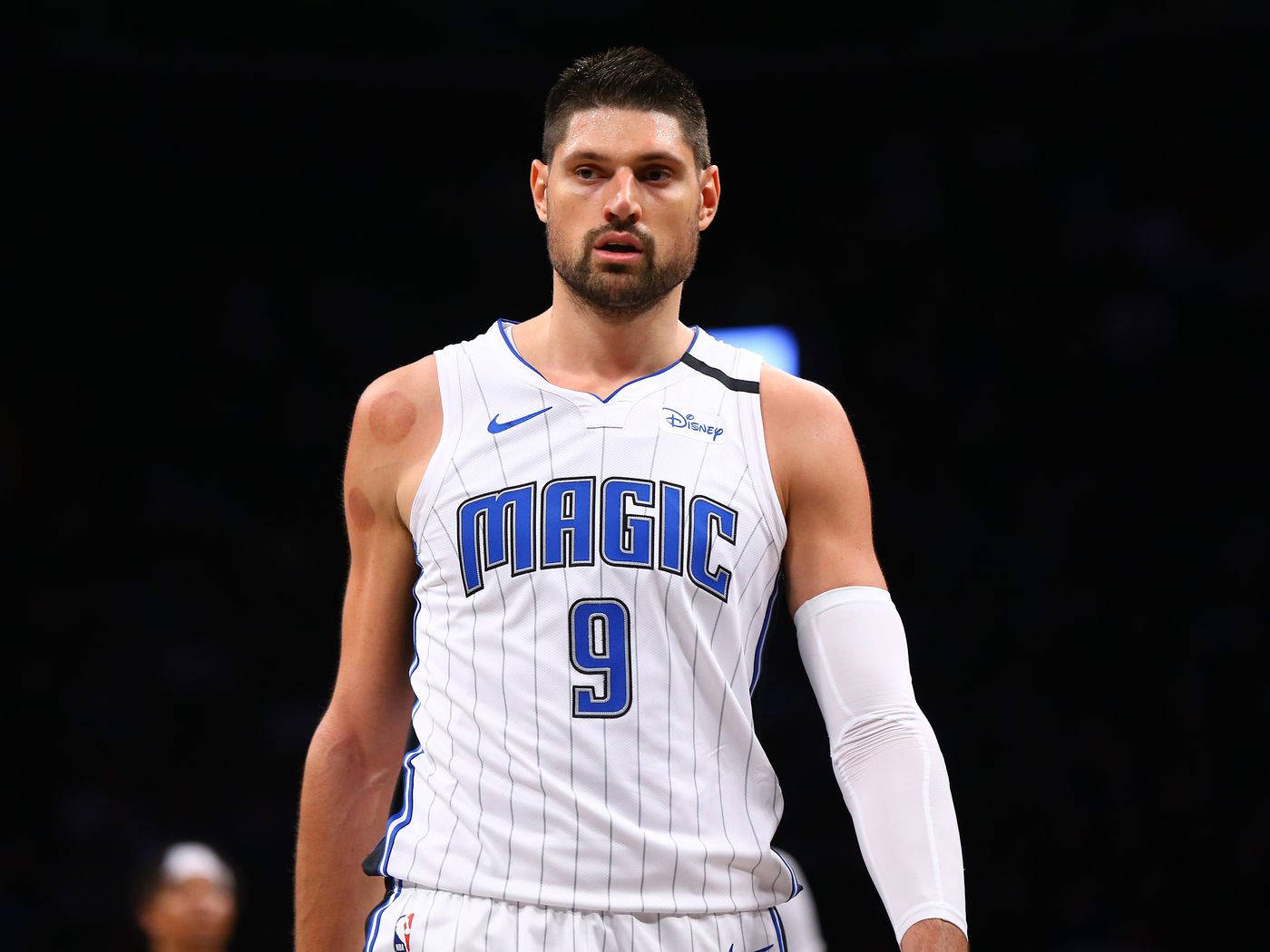 NBA球員 尼古拉·武切維奇 奧蘭多魔術隊 聚焦神情 壁紙 — 一位專注的籃球運動員，身穿帶有藍色飾邊的白色奧蘭多魔術隊球衣，站在黑暗的球場背景下。