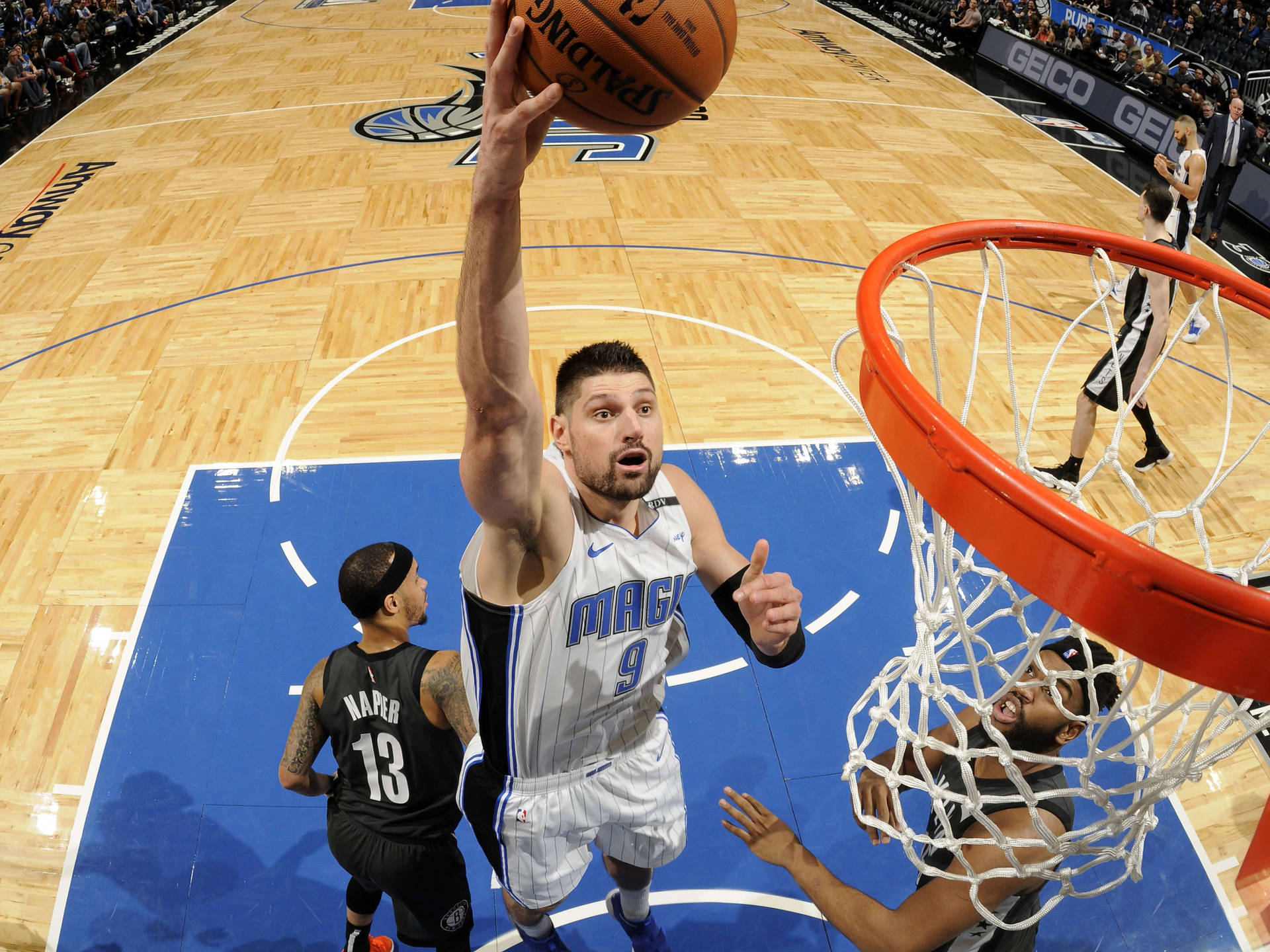 NBA球員 尼古拉·武切維奇 奧蘭多魔術隊 上籃投籃 壁紙 — 一名身穿魔術隊球衣的籃球運動員在防守球員從下方觀看時灌籃。