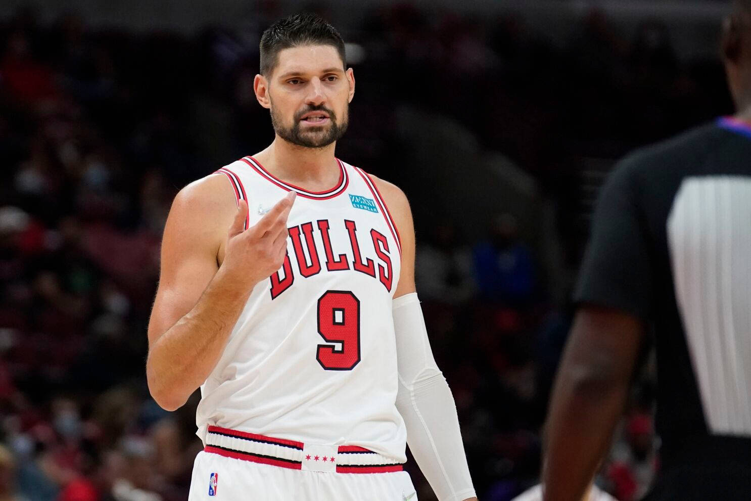 NBA球員 尼古拉·武切維奇 芝加哥公牛隊 與裁判對話 壁紙 — 一名專注的籃球運動員身穿白色公牛隊9號球衣，在明亮的場館燈光下比賽中做手勢。