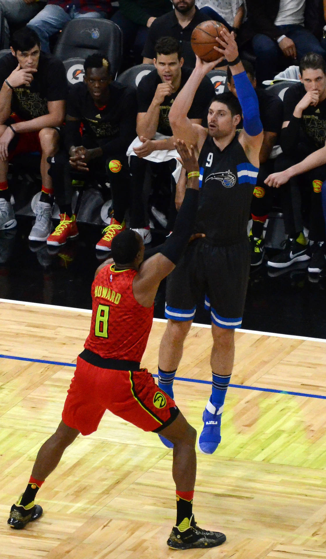 NBA球員奧蘭多魔術隊尼古拉·武切維奇投籃 壁紙 — 一名身穿黑色球衣的籃球運動員跳起投籃，而一名身穿紅色球衣的防守球員在明亮的場館燈光下試圖封阻投籃。