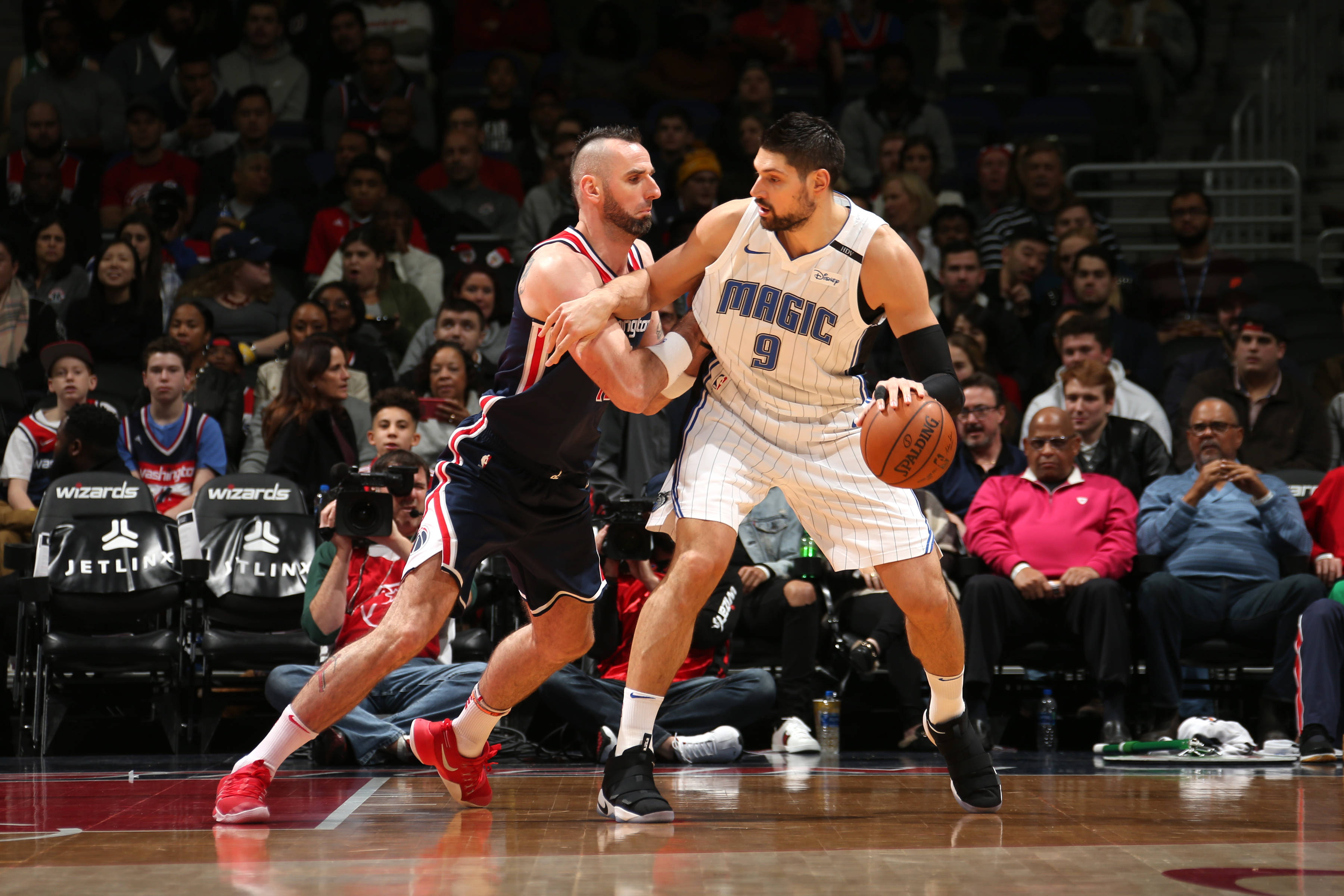 NBA球員奧蘭多魔術隊尼古拉·武切維奇防守 壁紙 — 一名身穿白色奧蘭多魔術隊球衣的籃球運動員在激烈的比賽中運球，同時被一名身穿黑色球衣的對手緊密防守。