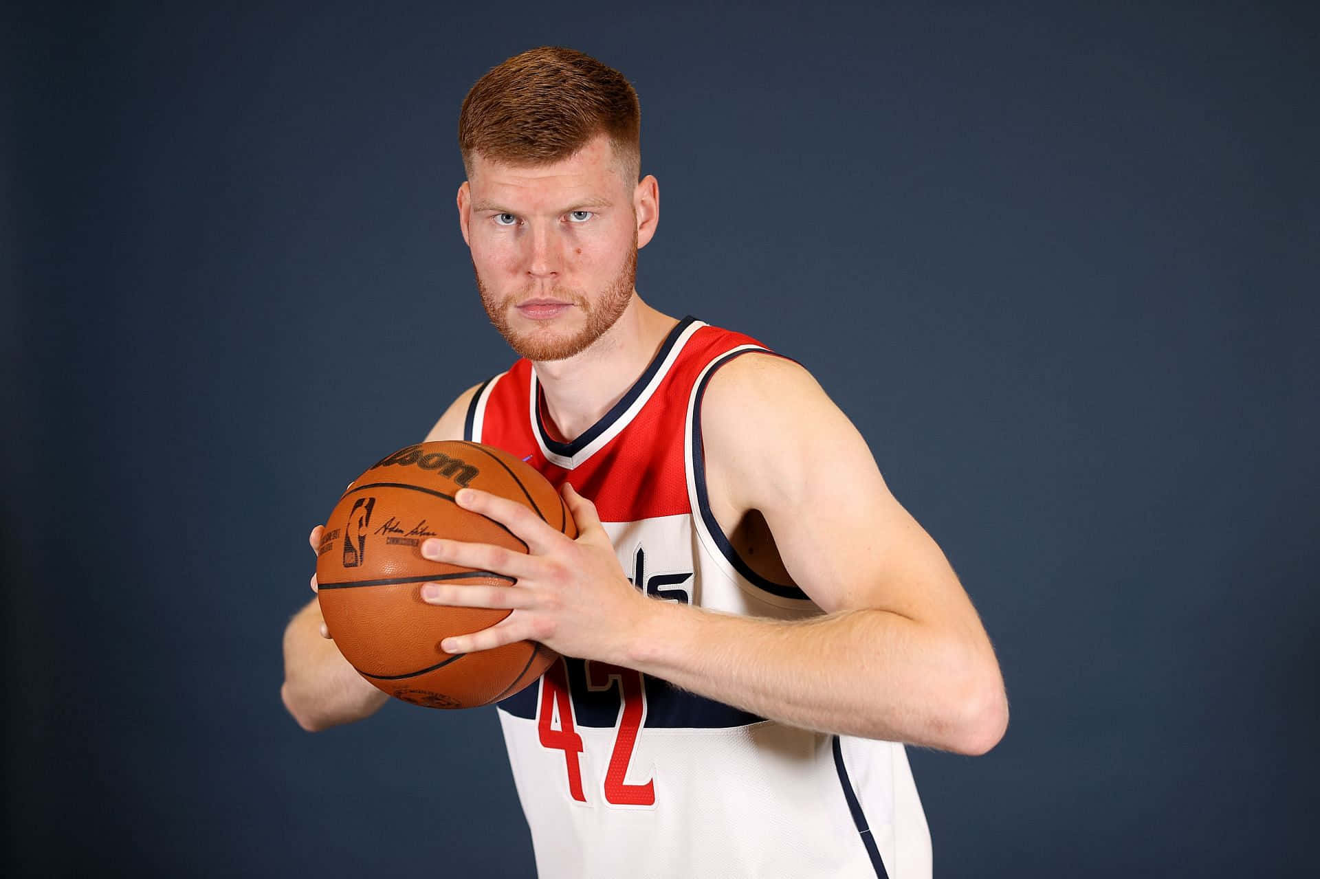 NBA球員戴維斯·貝坦斯肖像 壁紙 — 一位肌肉發達的籃球運動員身穿紅白球衣，在深藍色攝影棚背景下手持籃球。