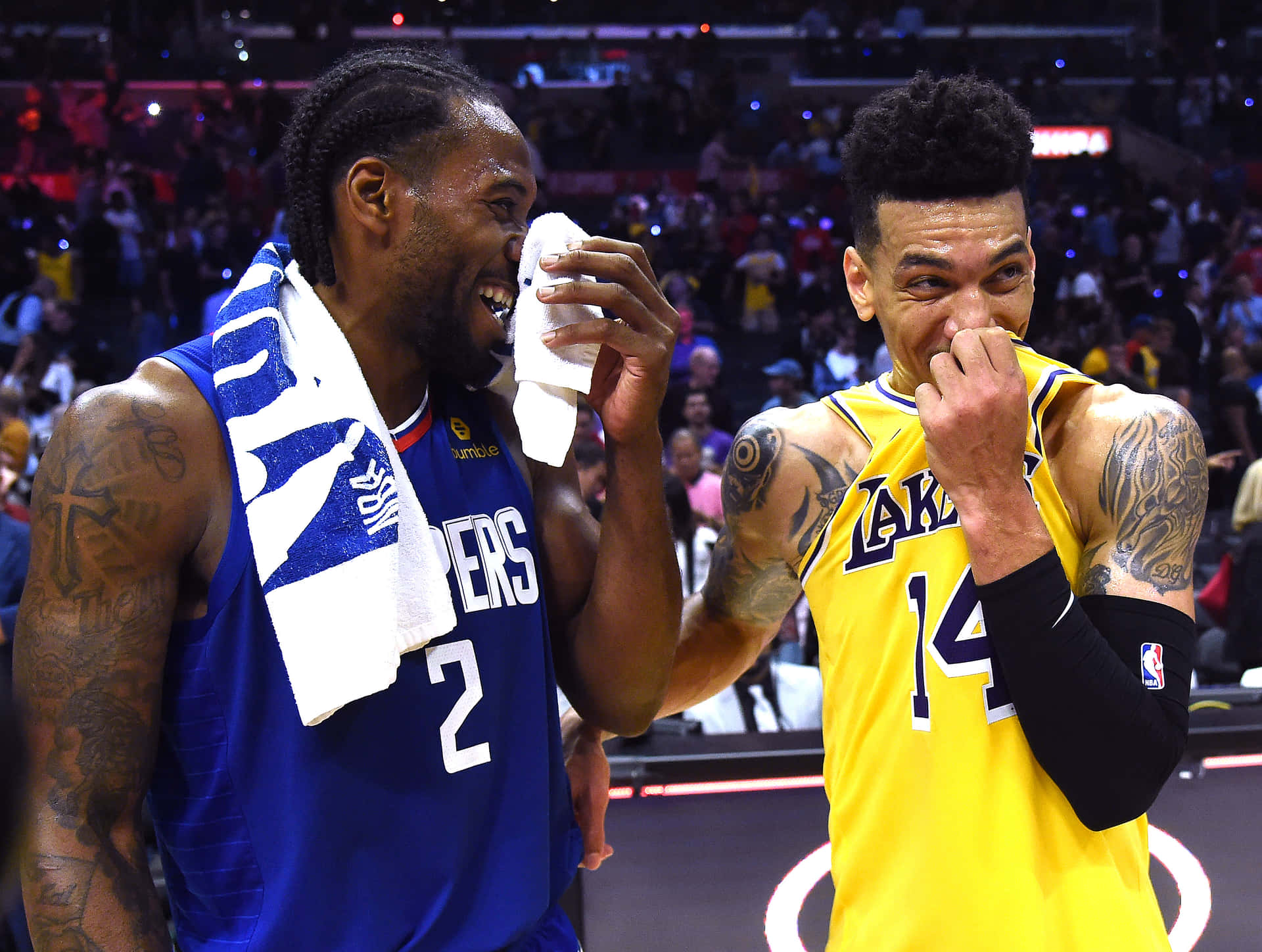 NBA球員搞笑大笑照片 壁紙 — 兩名身穿球隊球衣的籃球運動員笑著，在燈光明亮的球場上分享歡樂時刻，身後有歡呼的人群。