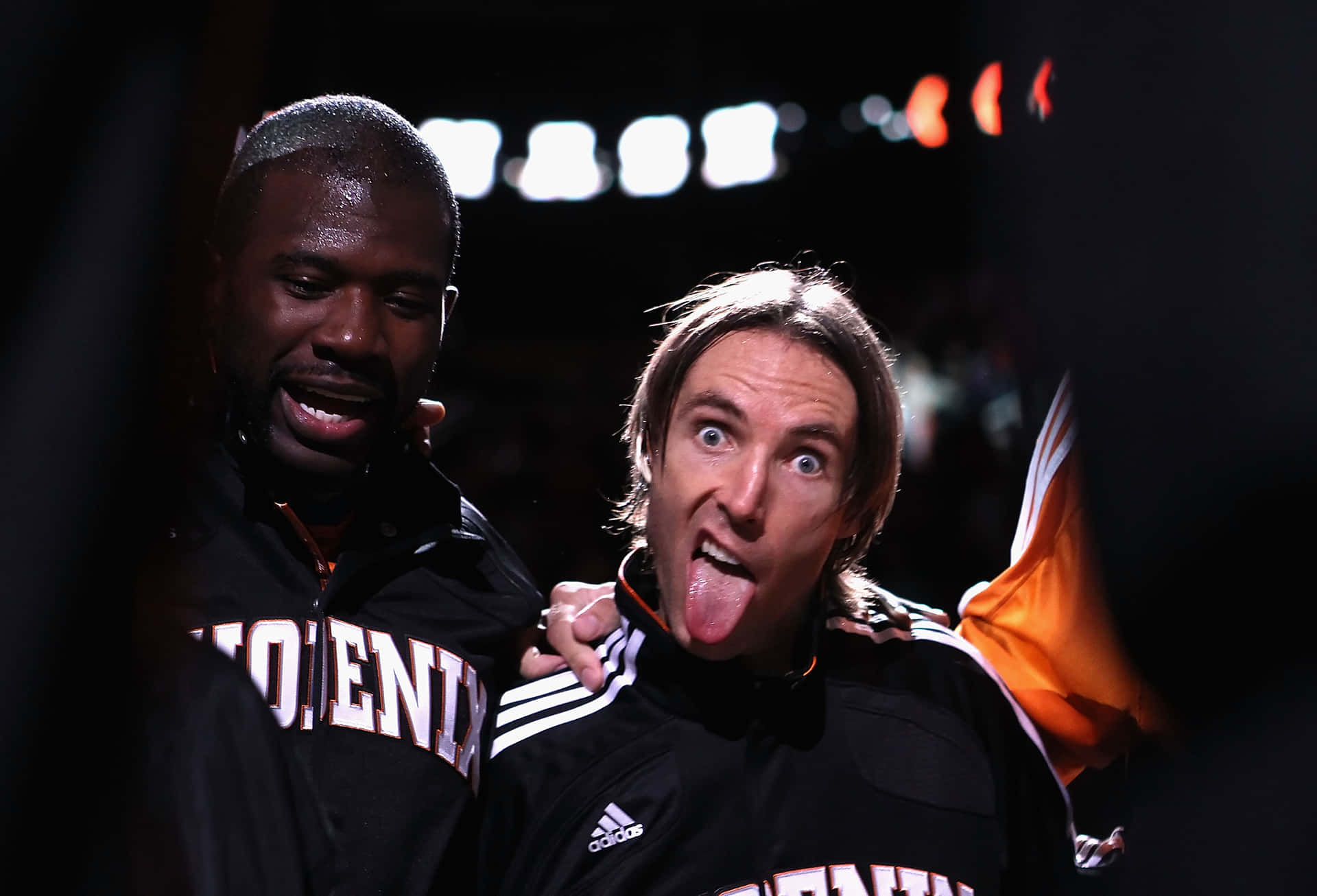 NBA球員搞笑姿勢照片 壁紙 — 兩名身穿鳳凰城太陽隊外套的籃球運動員在場館燈光下擺出俏皮姿勢，其中一人伸出舌頭。