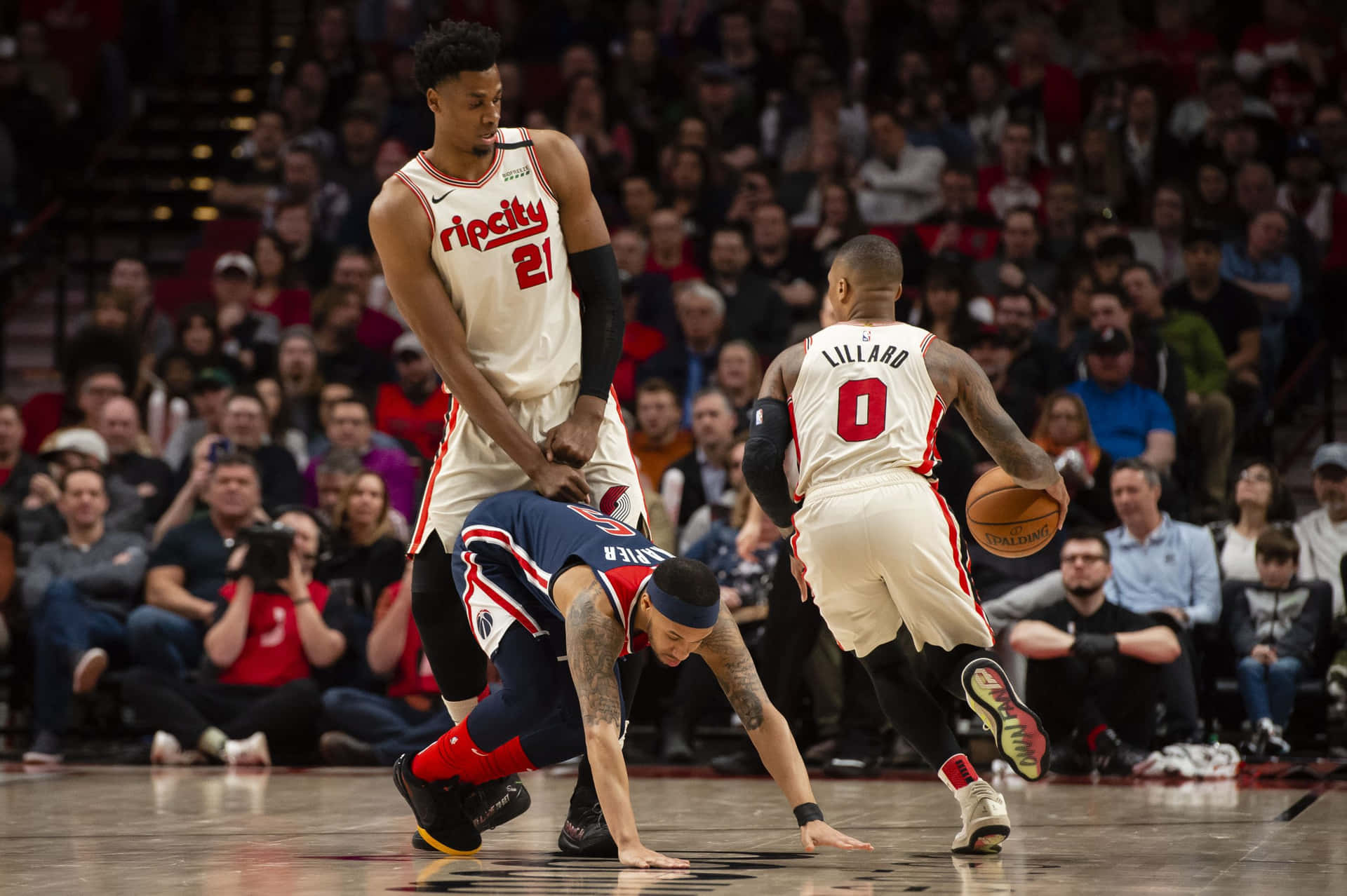 NBA球員搞笑姿勢照片 壁紙 — 一名身穿白色球衣的籃球運動員在擠滿觀眾的場館比賽中，運球越過倒地的對手，隊友則專注地觀看。