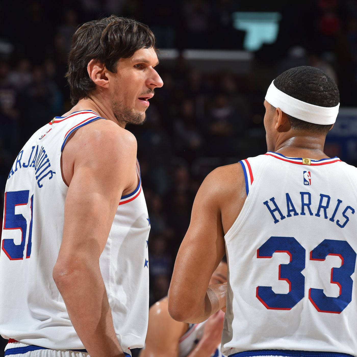 NBA球員經典搭檔托拜厄斯·哈里斯與博班·馬揚諾維奇 壁紙 — 兩名身穿白色球衣、帶有藍紅飾邊的籃球運動員在明亮的場館燈光下，於比賽中激烈對峙。