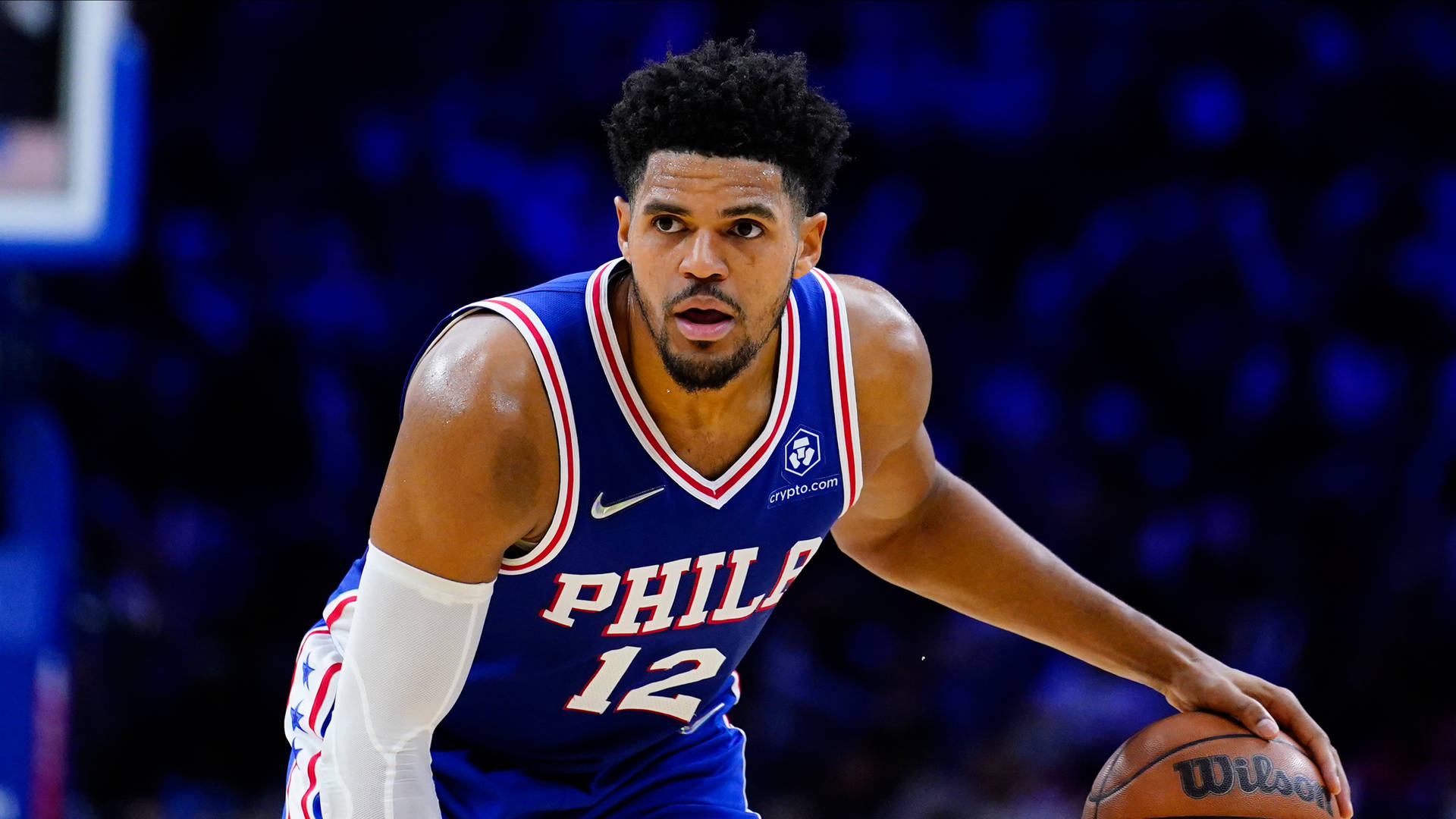 NBA球員費城76人隊托拜厄斯·哈里斯 壁紙 — 一位專注的籃球運動員身穿藍色費城球衣，在明亮的場館燈光下激烈運球。
