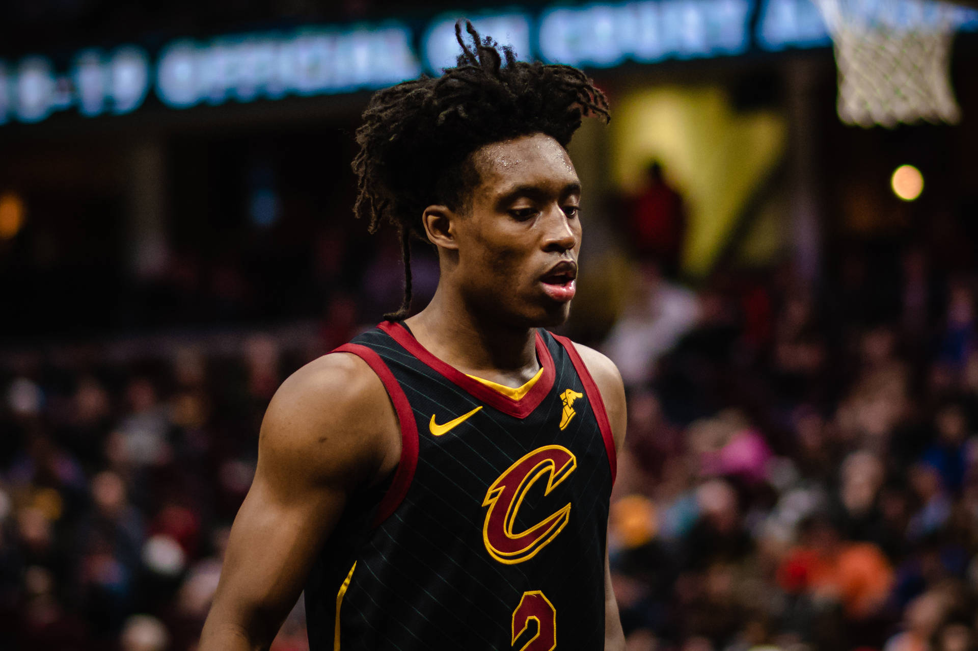 NBA球星柯林·塞克頓離場 壁紙 — 一位專注的籃球運動員身穿黑紅色的克里夫蘭騎士隊球衣，站在燈光明亮的球場上，背景是模糊的觀眾。