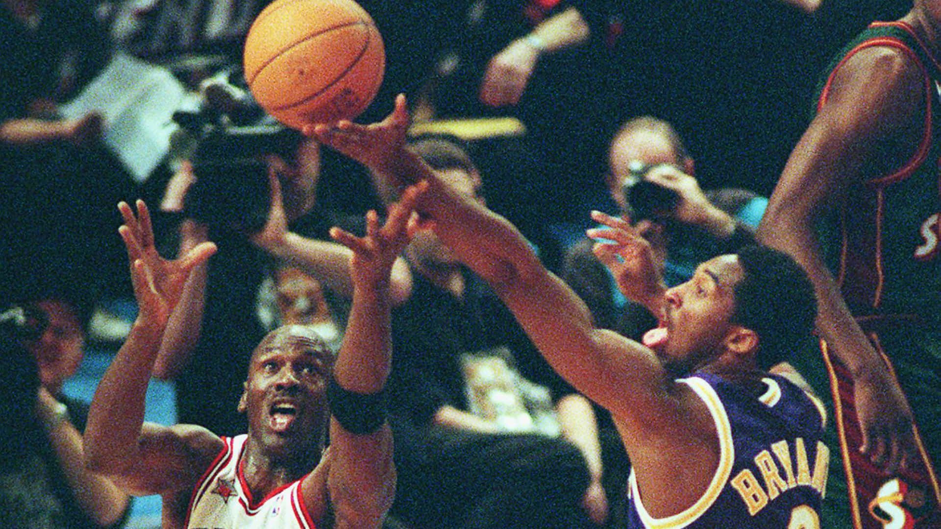 NBA球星科比·布萊恩特與邁克爾·喬丹全明星經典舊照 壁紙 — 米高佐敦跳起投籃，防守球員白賴仁梳爾在場館燈光下於緊張的籃球比賽中向上撲去。