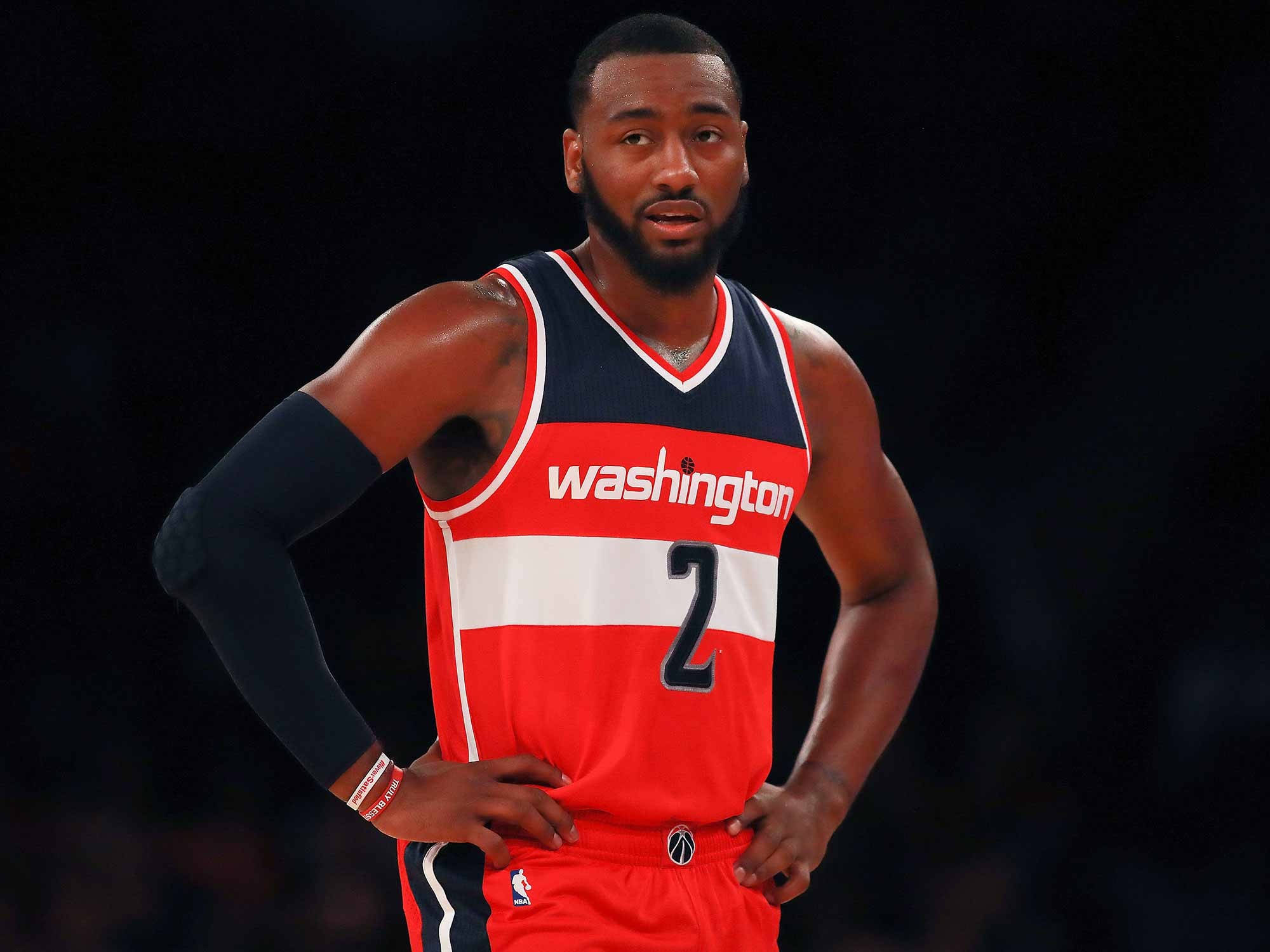 NBA球星約翰·沃爾帥氣風采 壁紙 — 一位專注的籃球運動員身穿紅白藍華盛頓球衣，背號二號，雙手叉腰站在黑暗的球場背景下。