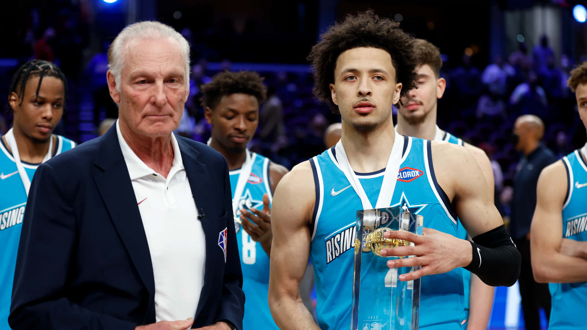 NBA瑞克·巴里與凱德·庫明加 壁紙 — 一名身穿藍色Rising Stars球衣的年輕籃球運動員，在燈光璀璨的場館中，手持獎盃，站在一位身穿西裝的年長男子旁邊。