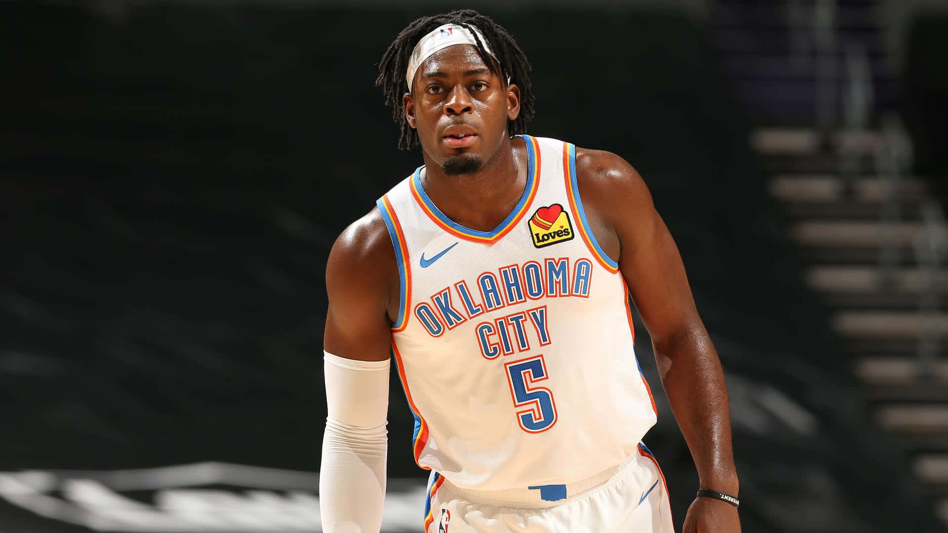 NBA盧根茲·多特5號球衣 壁紙 — 一名專注的籃球運動員身穿奧克拉荷馬城球衣，在燈光昏暗的球場上準備就緒，身後是模糊的體育館座位。