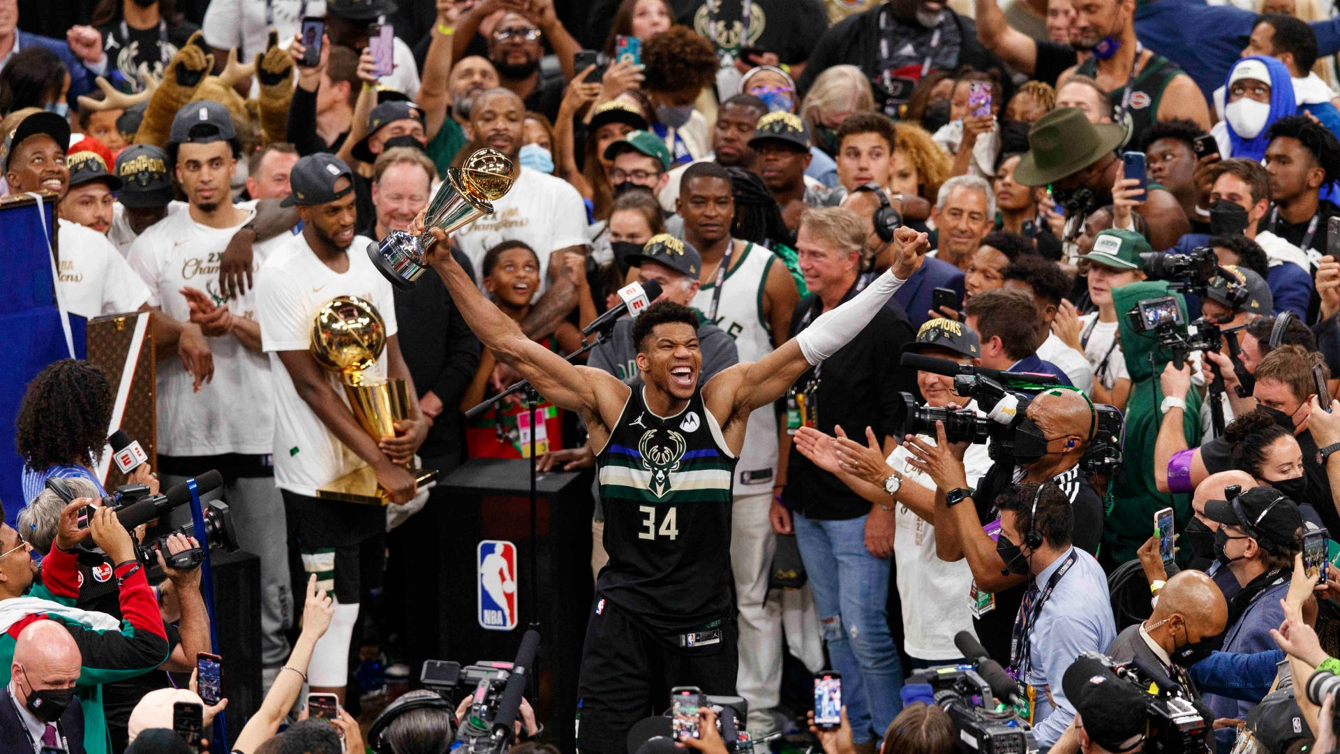 NBA總決賽 吉安尼斯·安特托昆博 密爾沃基冠軍 壁紙 — 一名身穿黑色球衣、背號34的籃球運動員在歡呼的球迷和媒體包圍下，勝利地將金色獎盃高舉過頭頂。
