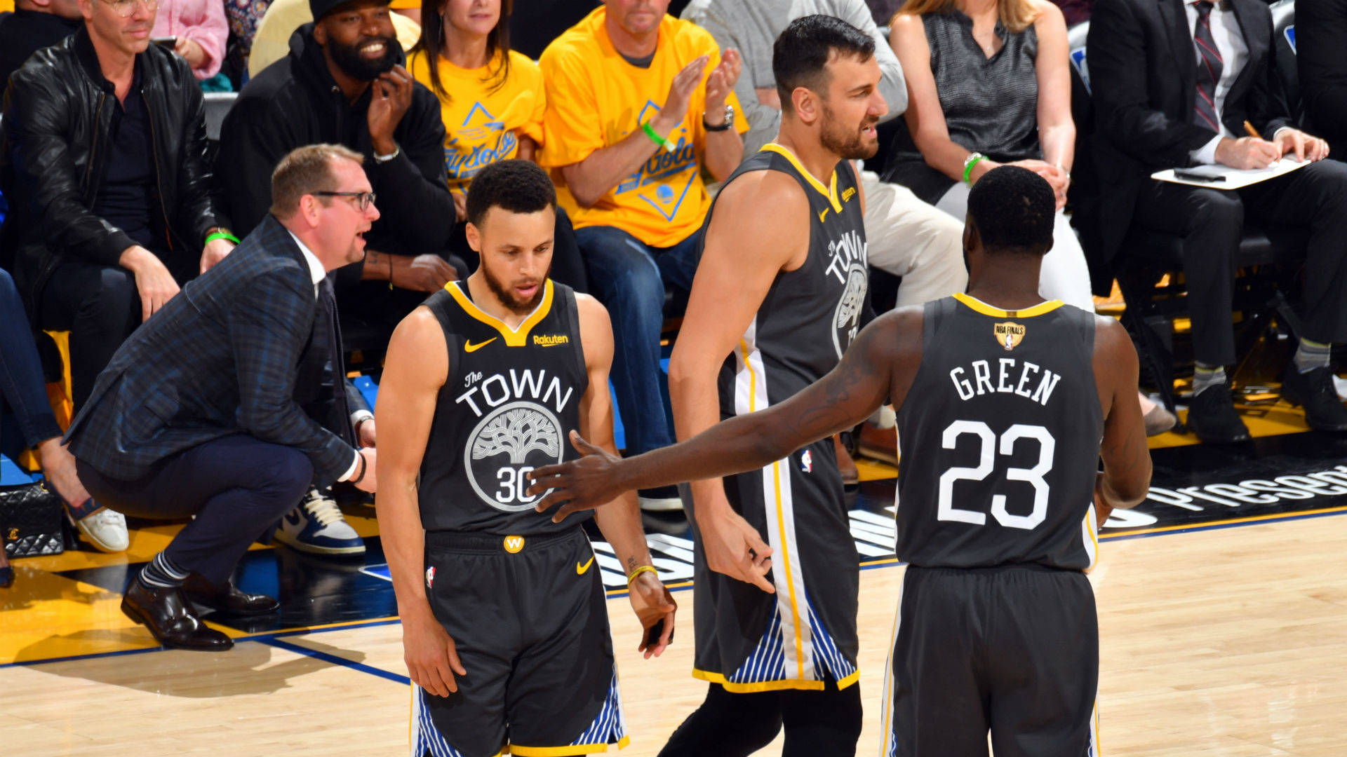 NBA總決賽勇士隊比賽 壁紙 — 金州勇士隊球員身穿黑色和黃色球衣，在光亮的籃球場上比賽時互動，背景有觀眾。