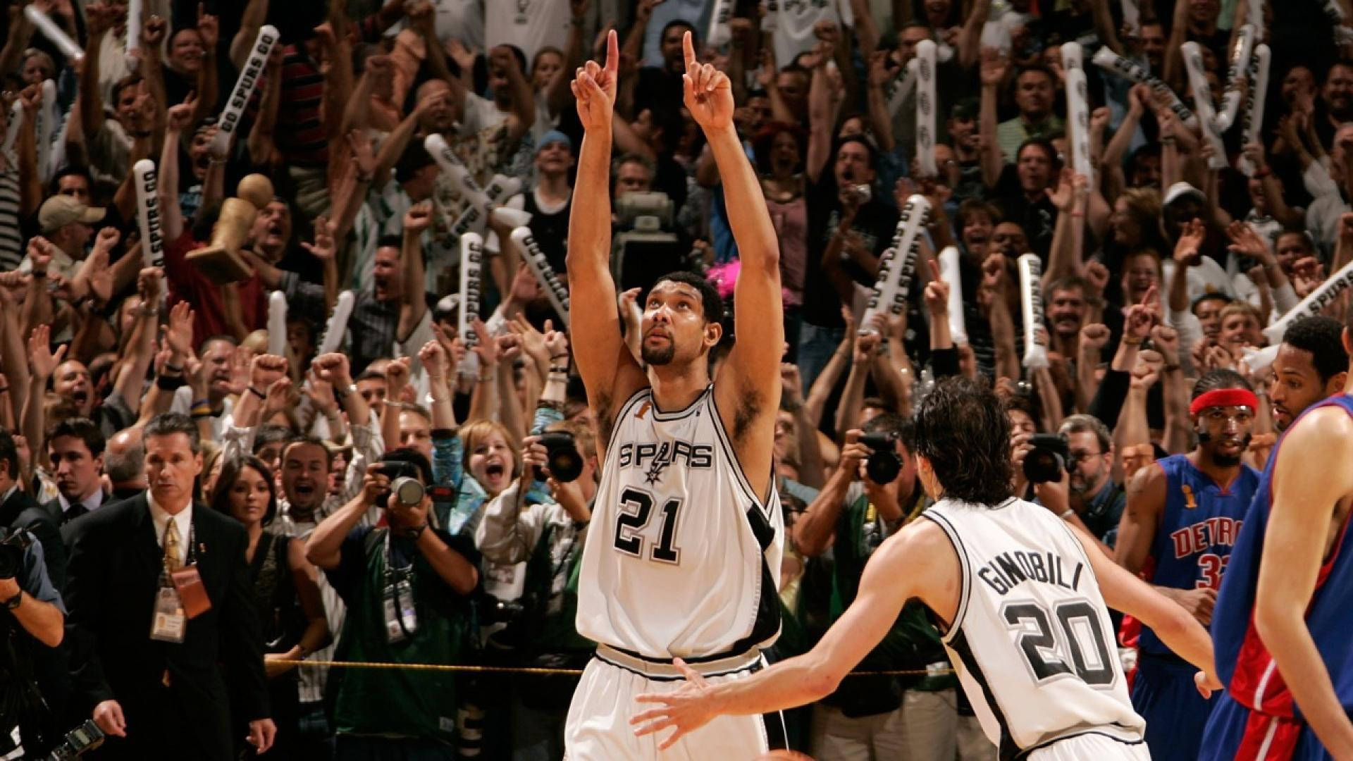 NBA總決賽精彩瞬間 壁紙 — 一名身穿馬刺隊球衣的籃球運動員勝利地舉起雙食指，看台上的球迷揮舞著泡沫手指瘋狂歡呼。
