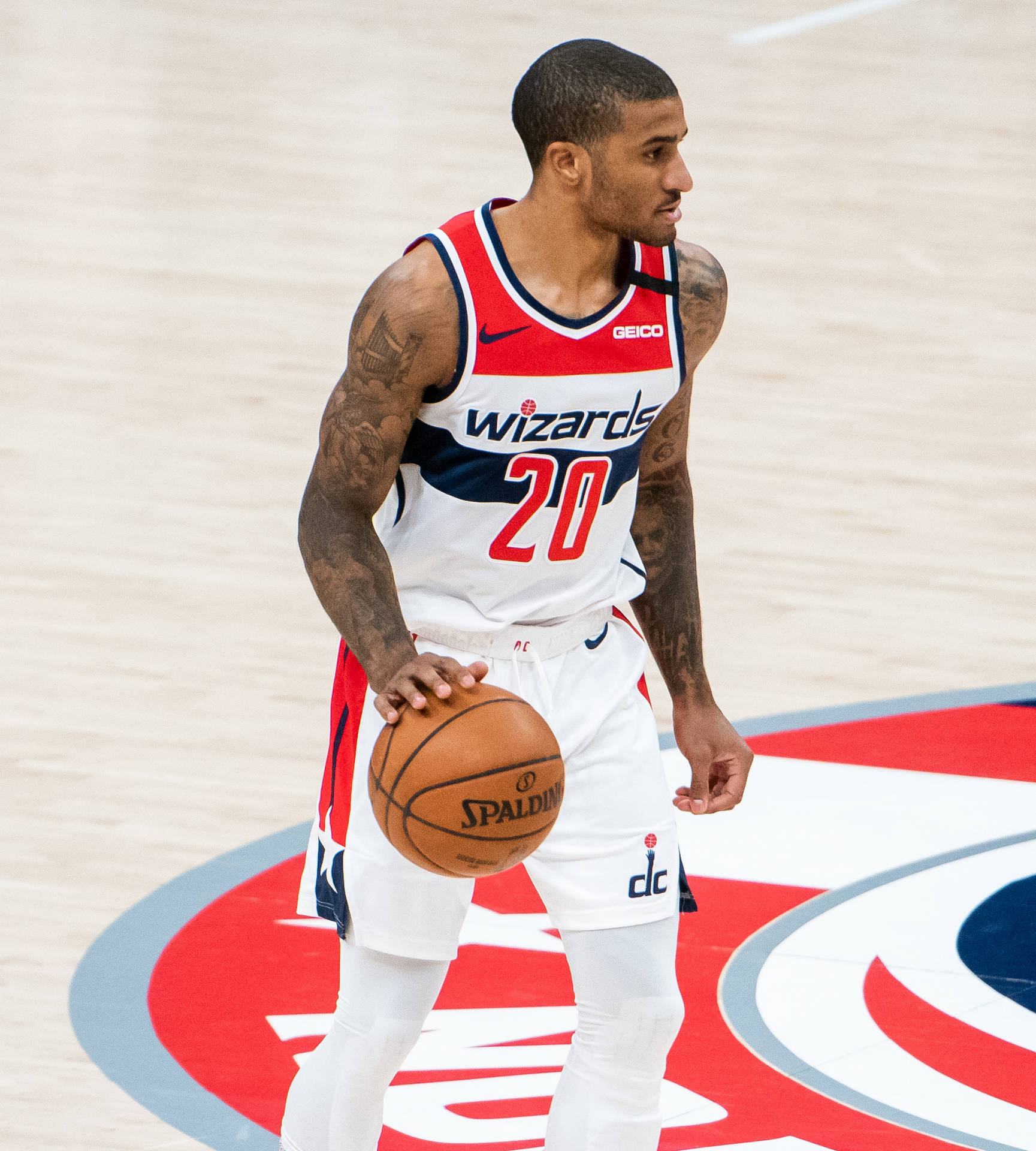 NBA華盛頓巫師隊加里·佩頓二世 壁紙 — 一名身穿巫師隊球衣的籃球運動員，在明亮的場館燈光下，於帶有球隊標誌的球場上手持 Spalding 籃球。