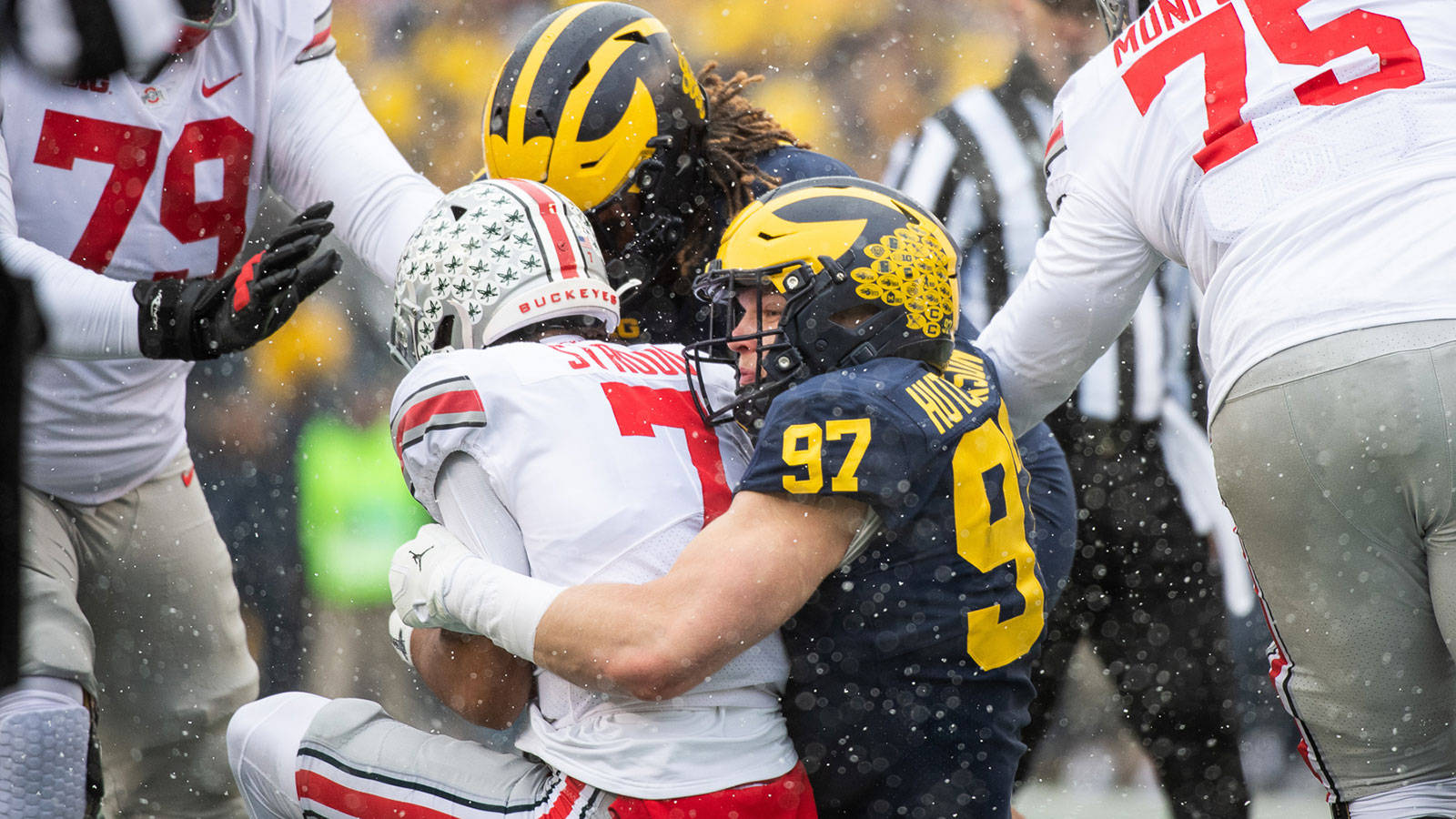 NCAA Aidan Hutchinson 密歇根對俄亥俄州立大學對抗賽 壁紙 — 身穿白色和海軍藍色球衣的足球運動員在雪地比賽中激烈纏鬥，雪花在他們周圍飄落。
