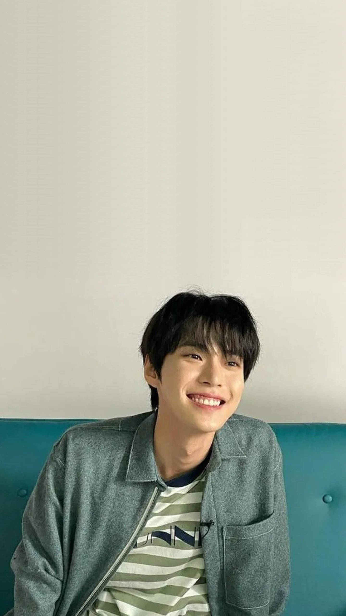 NCT 127 Doyoung 的甜美笑容 壁紙 — 一位深色頭髮的年輕男子坐在青綠色簇絨沙發上，對著白色牆壁露出溫暖的笑容。