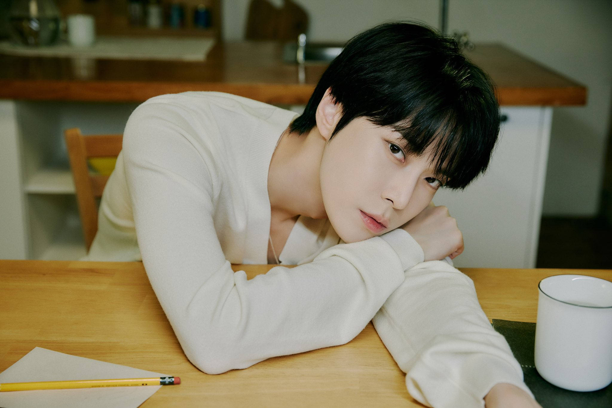 NCT 127 Doyoung「飛毯之旅」預告照 壁紙 — 一位深色頭髮的年輕男子在柔和光線的廚房裡，將頭靠在木桌上的手臂上。