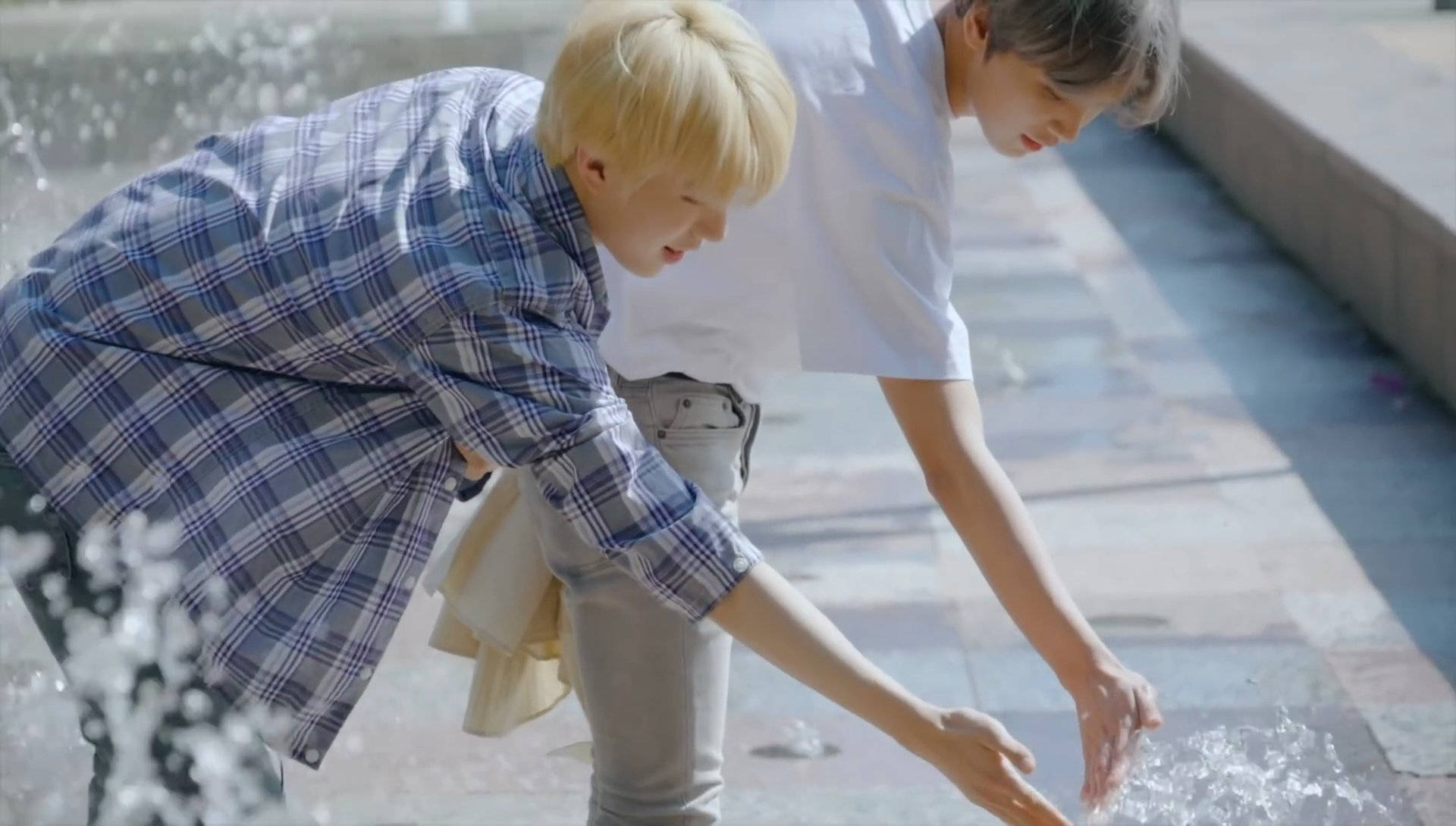 NCT 127 Jungwoo 與 Haechan 玩水歡樂時光 壁紙 — 兩名身穿休閒服的年輕男子在陽光普照的瓷磚小徑上嬉戲地潑水。