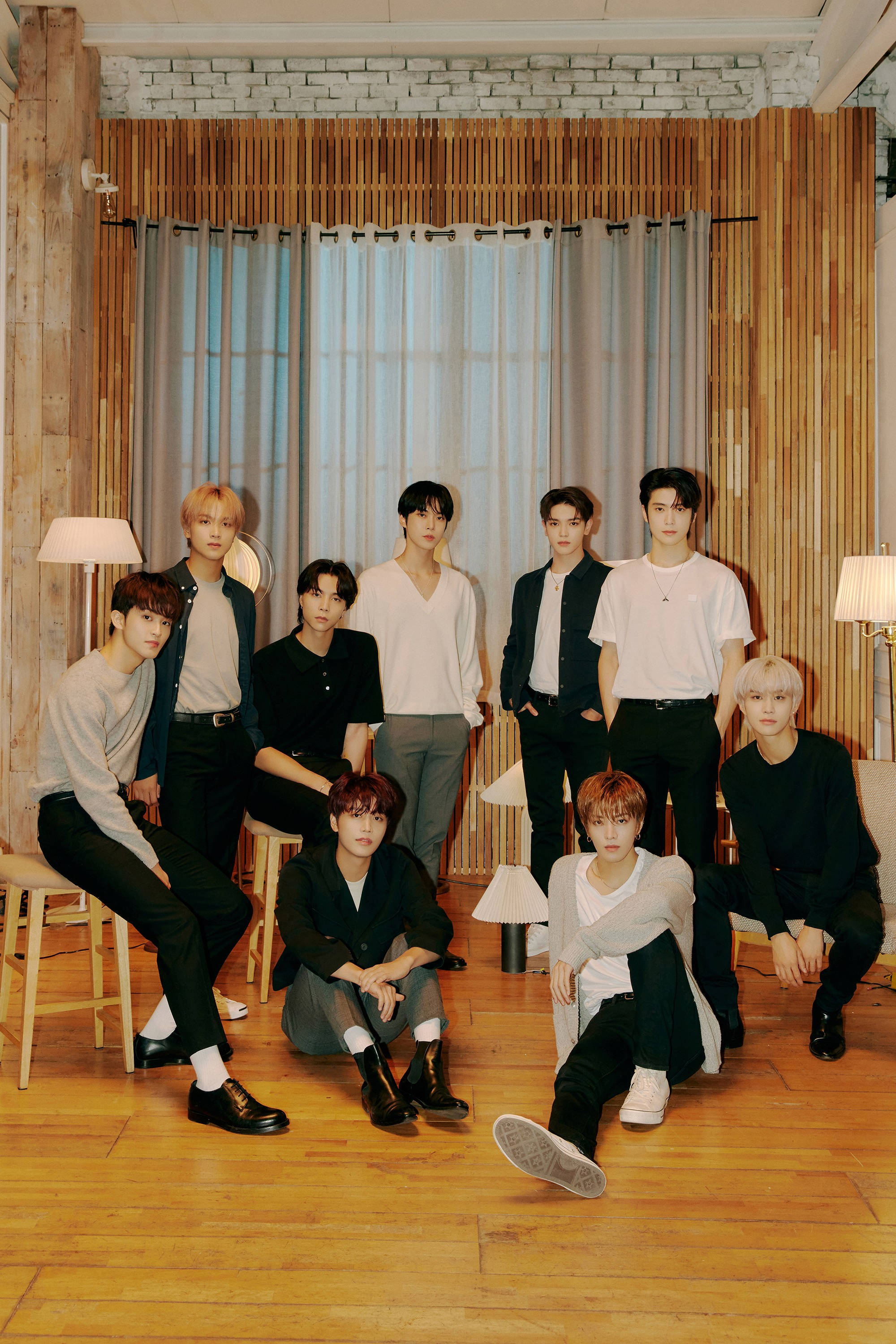NCT 127《Magic Carpet Ride》MV 預告照 壁紙 — 九位時尚年輕男士在一個現代木質房間內隨意擺姿勢，房間有磚牆和落地燈。
