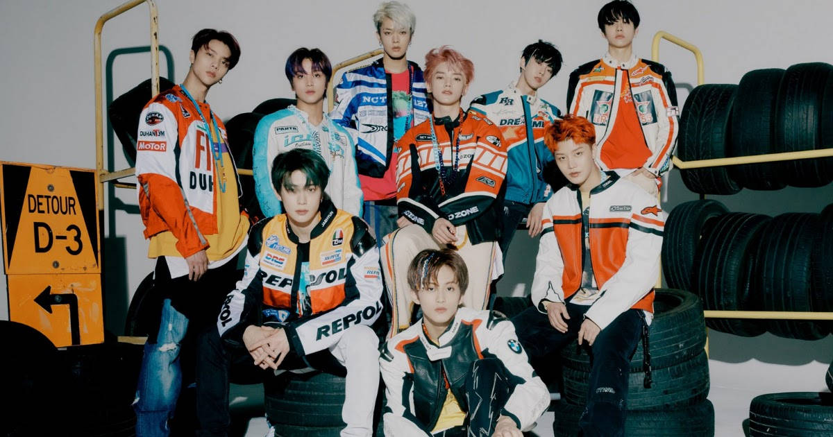 NCT 127 Neo Zone 預告影像 壁紙 — 七位身穿色彩繽紛賽車服的年輕男子在車庫環境中，自信地擺姿勢，周圍堆疊著輪胎和一個繞道標誌。