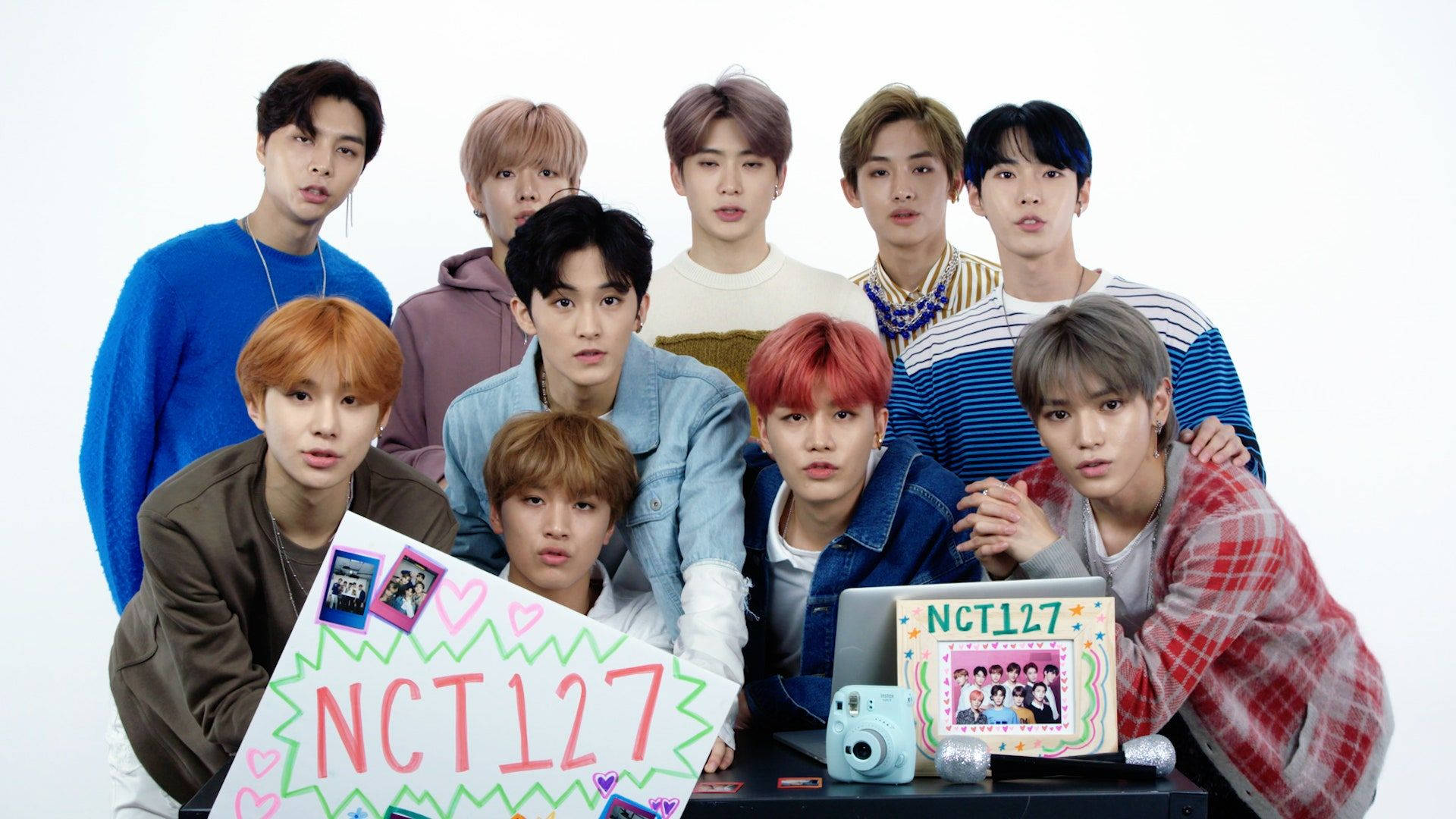 NCT 127 Q&A 現場即拍照 壁紙 — 九位年輕男子身穿色彩繽紛的休閒服裝，手持NCT127標語牌和相機，在純白背景前一起擺姿勢。