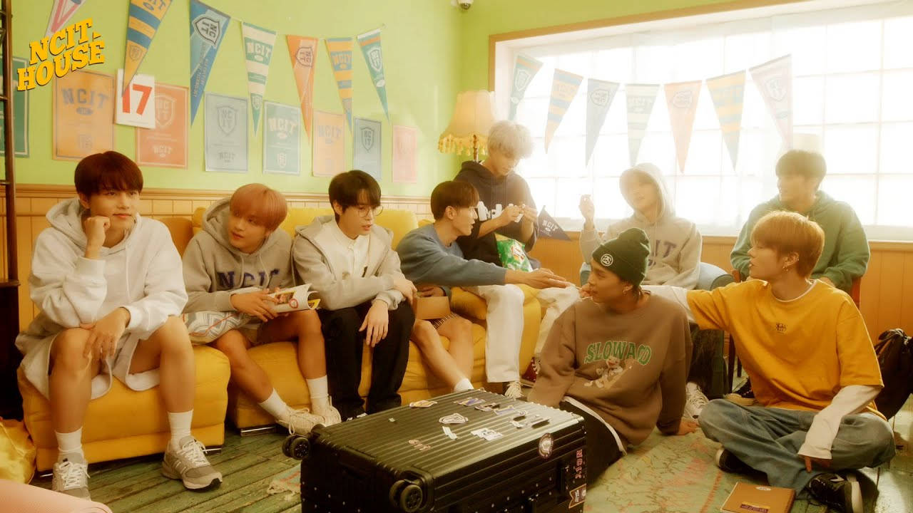 NCT 127 在 NCIT House 2021 壁紙 — 九個穿著休閒服的年輕男子放鬆地坐在黃色梳化和地板上，房間光線充足，裝飾著彩色三角旗。