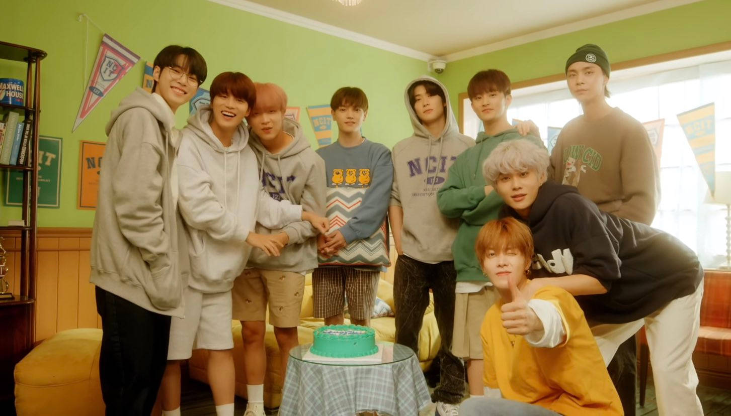 NCT 127 在 NCIT House 2021 壁紙 — 七位年輕男性穿著休閒連帽衫和運動褲，在一個光線充足、有綠色牆壁和體育橫幅的房間裡，圍著一個小綠色蛋糕愉快地擺姿勢。