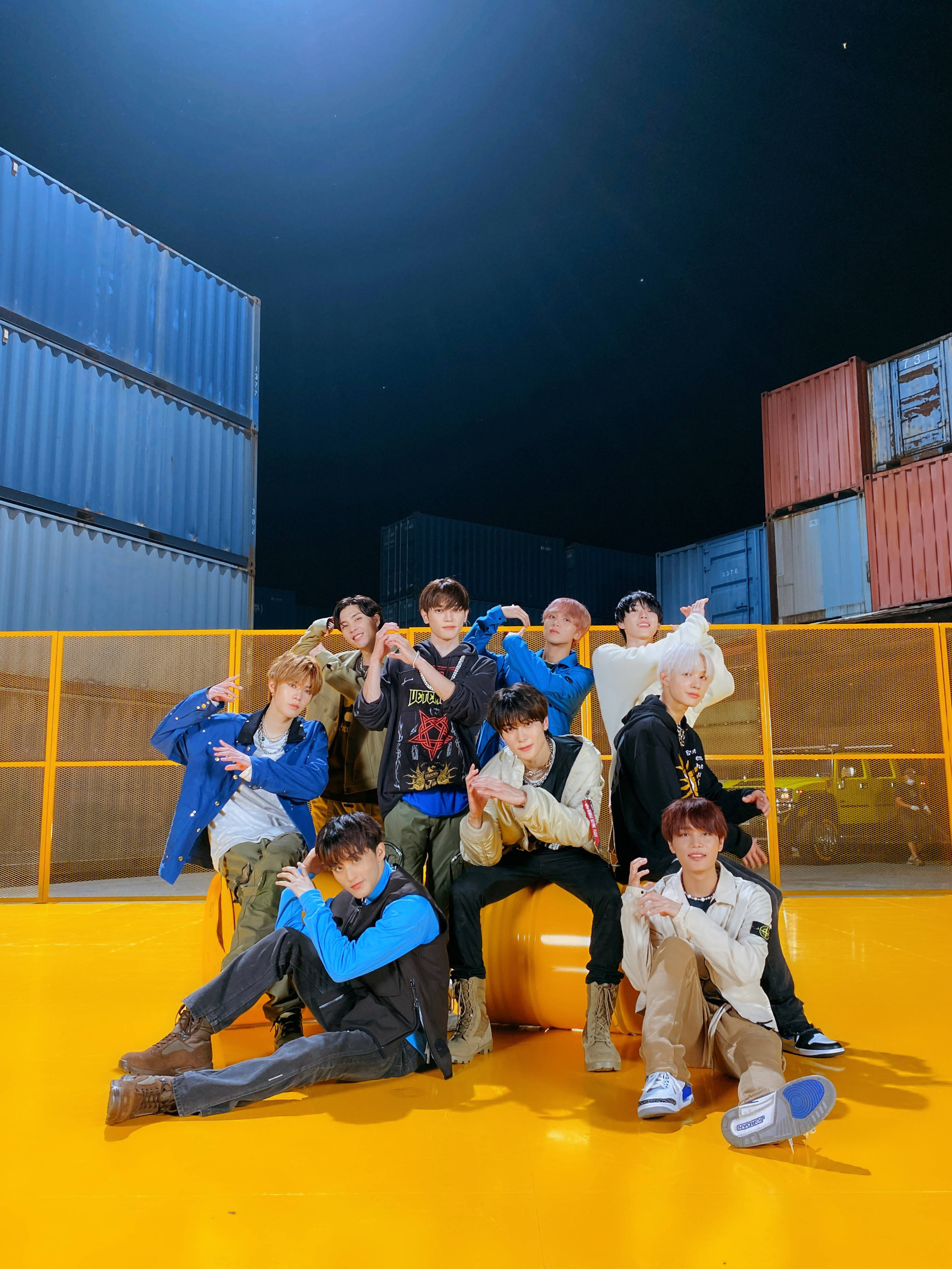 NCT 127 檸檬水姿勢 壁紙 — 九名年輕男子在明亮的黃色地板上自信地擺姿勢，背景是堆疊的貨運集裝箱，上方是繁星點點的夜空。