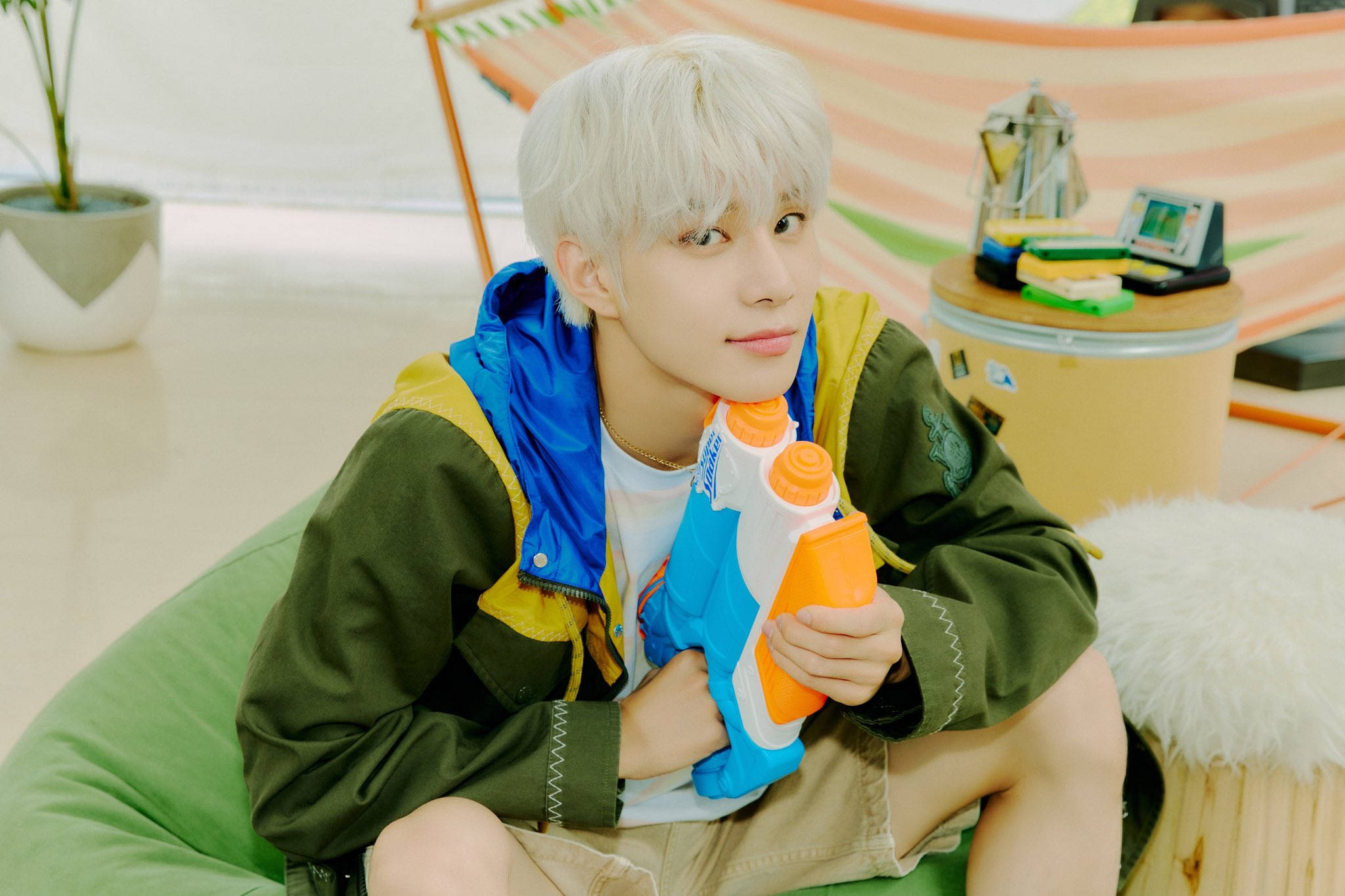 NCT 127 洪知秀《Road Trip》預告 壁紙 — 一位年輕人有鉑金色頭髮，坐在綠色豆袋椅上，拿著藍色和橙色的水槍，在明亮的室內空間。