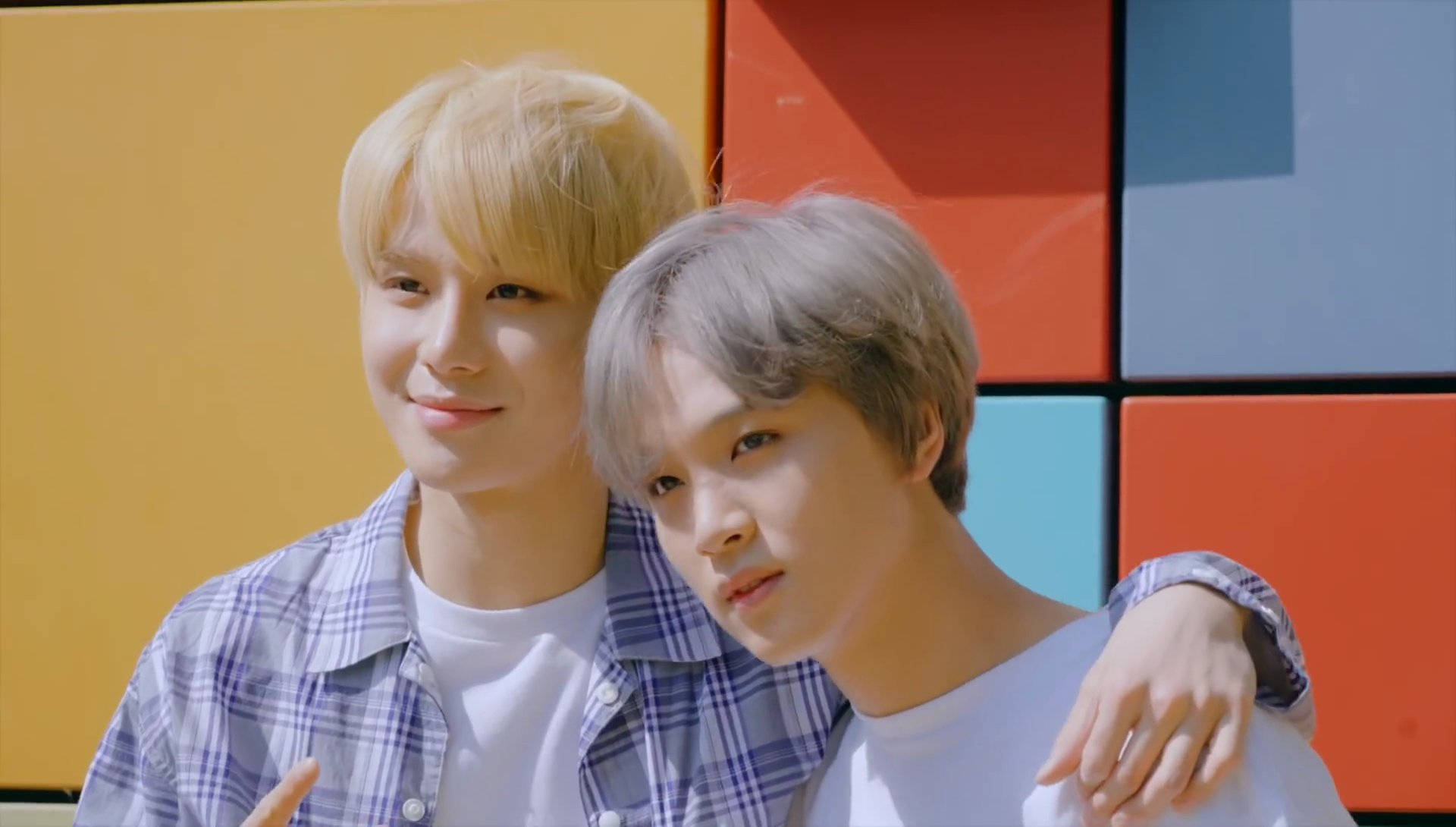 NCT 127 洪知秀與楷燦 壁紙 — 兩位年輕男子，一頭金髮一頭灰髮，緊靠著色彩繽紛的幾何牆壁，一人溫暖微笑，另一人若有所思地凝視。