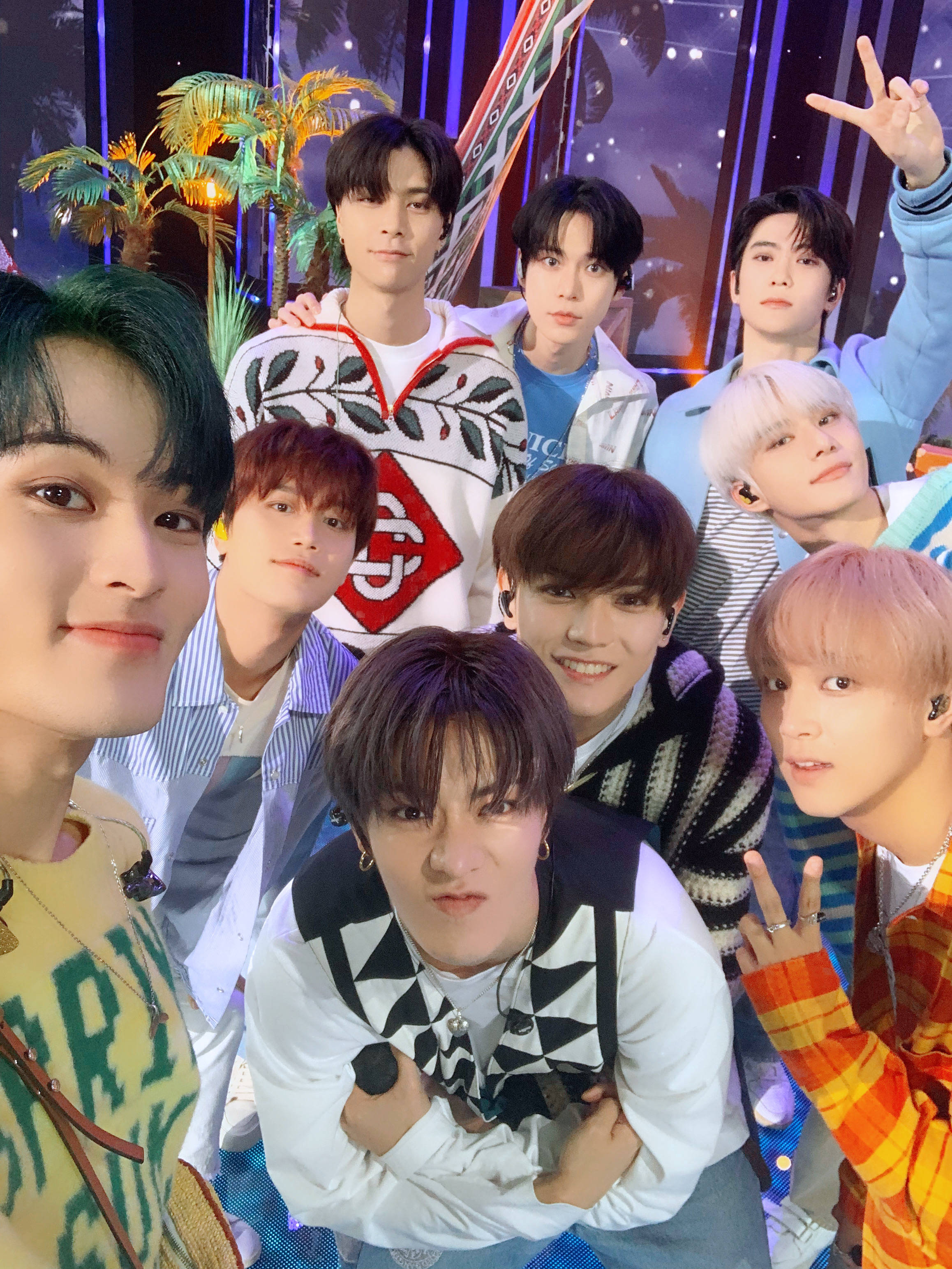 NCT 127 紀念公路之旅 壁紙 — 七位年輕男子，頭髮色彩繽紛，穿著時尚，在充滿活力的舞台場景中，棕櫚樹和閃爍燈光下愉快地擺姿勢。