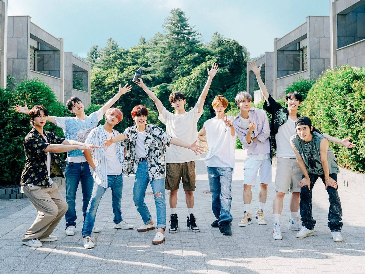 NCT 127《NCT Life》節目照 壁紙 — 九名年輕男子身穿休閒服裝，在陽光明媚的鋪砌庭院中擺出動態姿勢，周圍環繞著綠化和現代建築。
