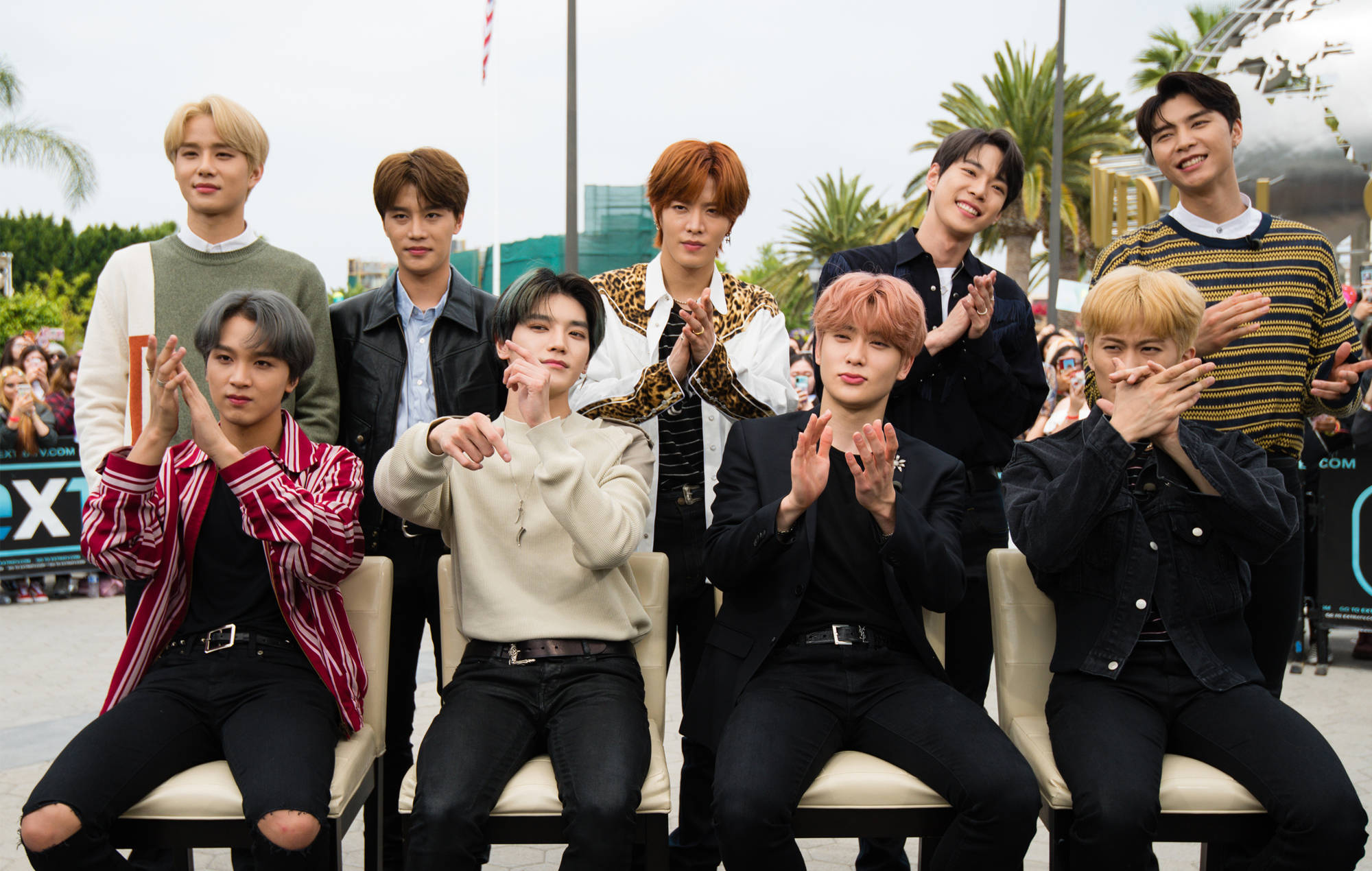NCT 127於好萊塢環球影城 壁紙 — 九位時尚年輕男士穿著潮流服裝，在戶外與棕櫚樹和身後的人群一起愉快地擺姿勢。