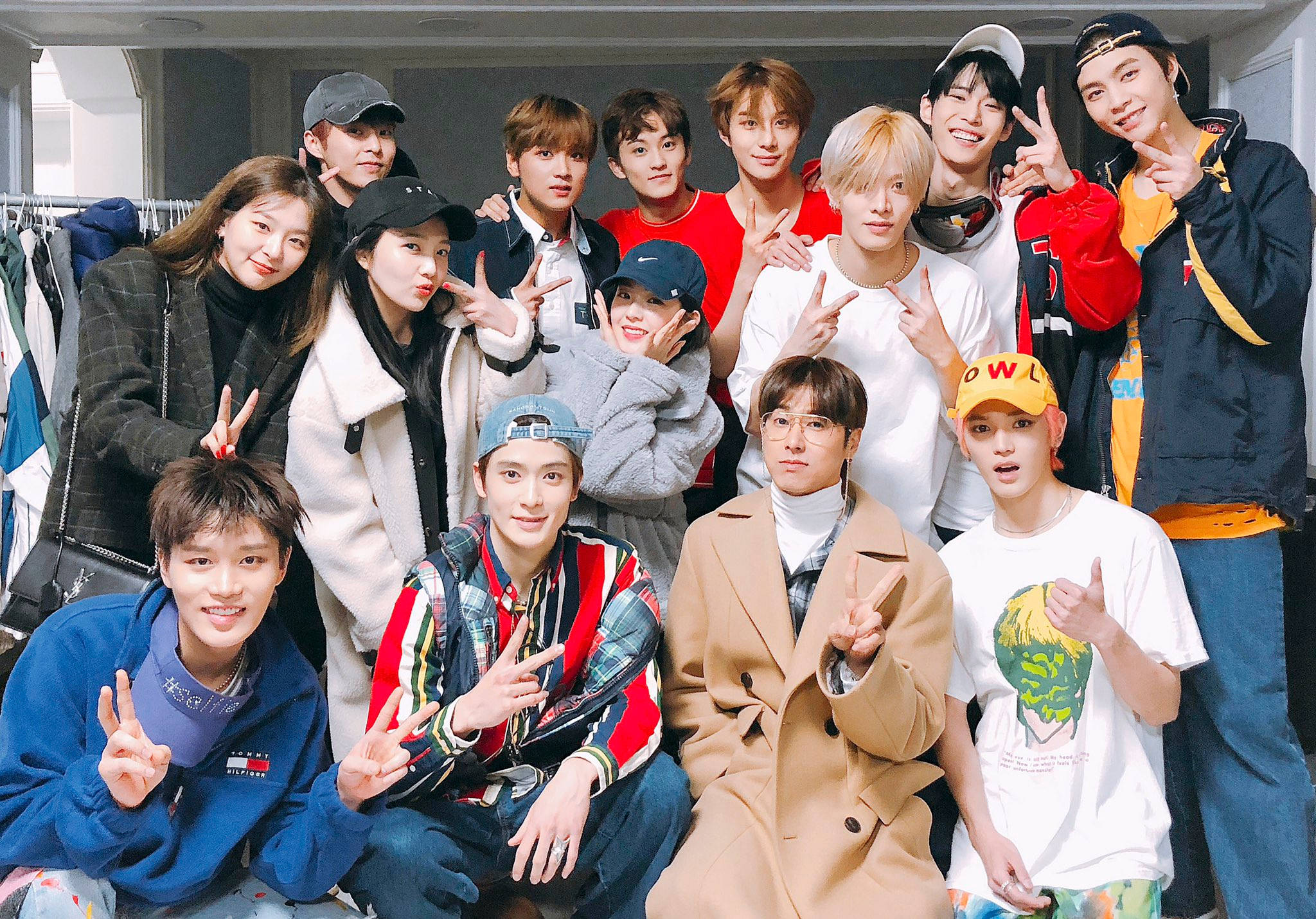 NCT 127與SM藝人聯合演唱會 壁紙 — 一群充滿活力的年輕人在服裝店裡一起擺出和平手勢，在自然光下燦爛微笑。