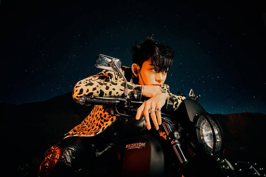 NCT Lucas 騎摩托車 壁紙 — 一名穿著豹紋外套的年輕男子在星空下靠著電單車，背景是黑暗的山脈。