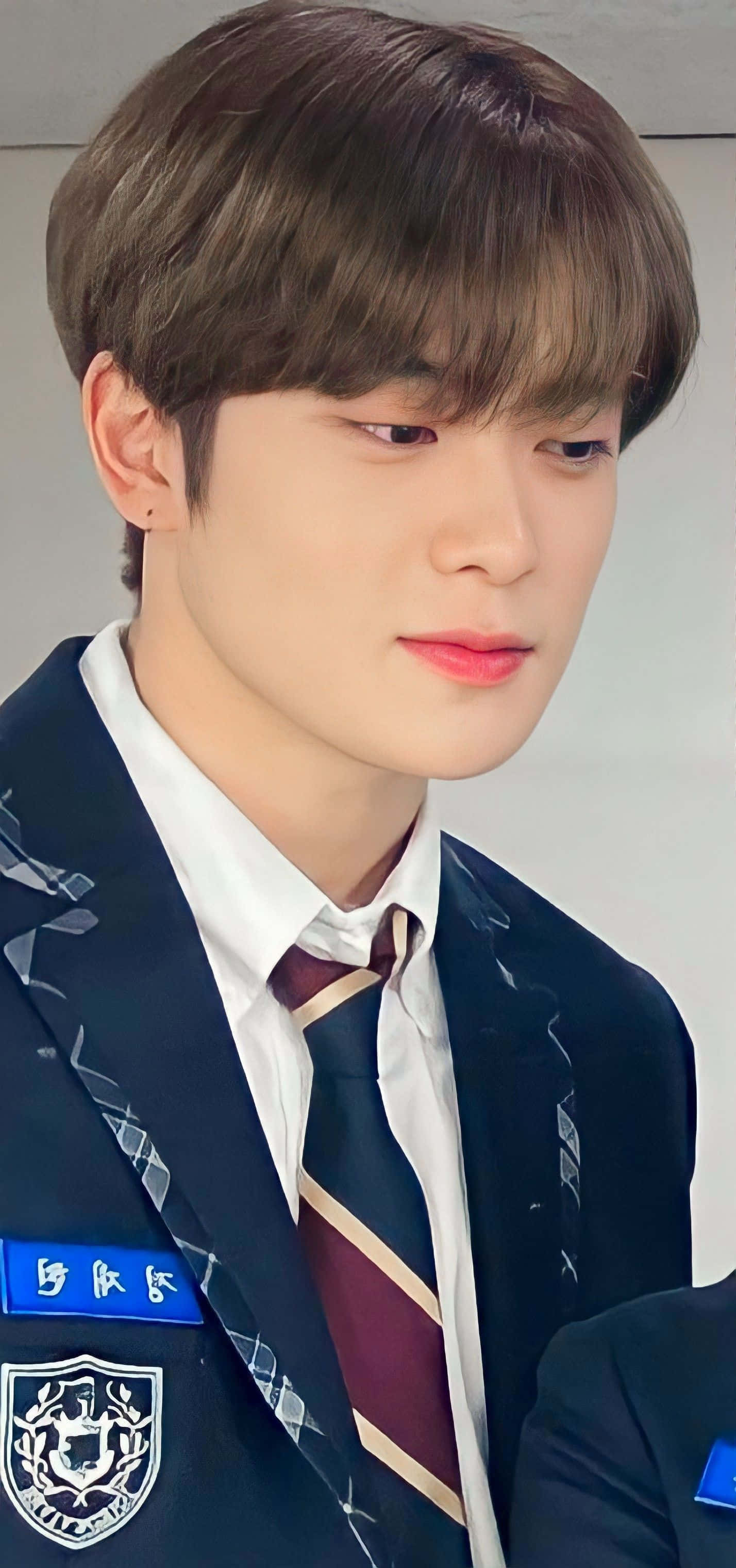 NCT 金廷祐 穿著經典校服 壁紙 — 一名身穿深色校服、打著條紋領帶的年輕男子，在樸素背景下若有所思地向下凝視。