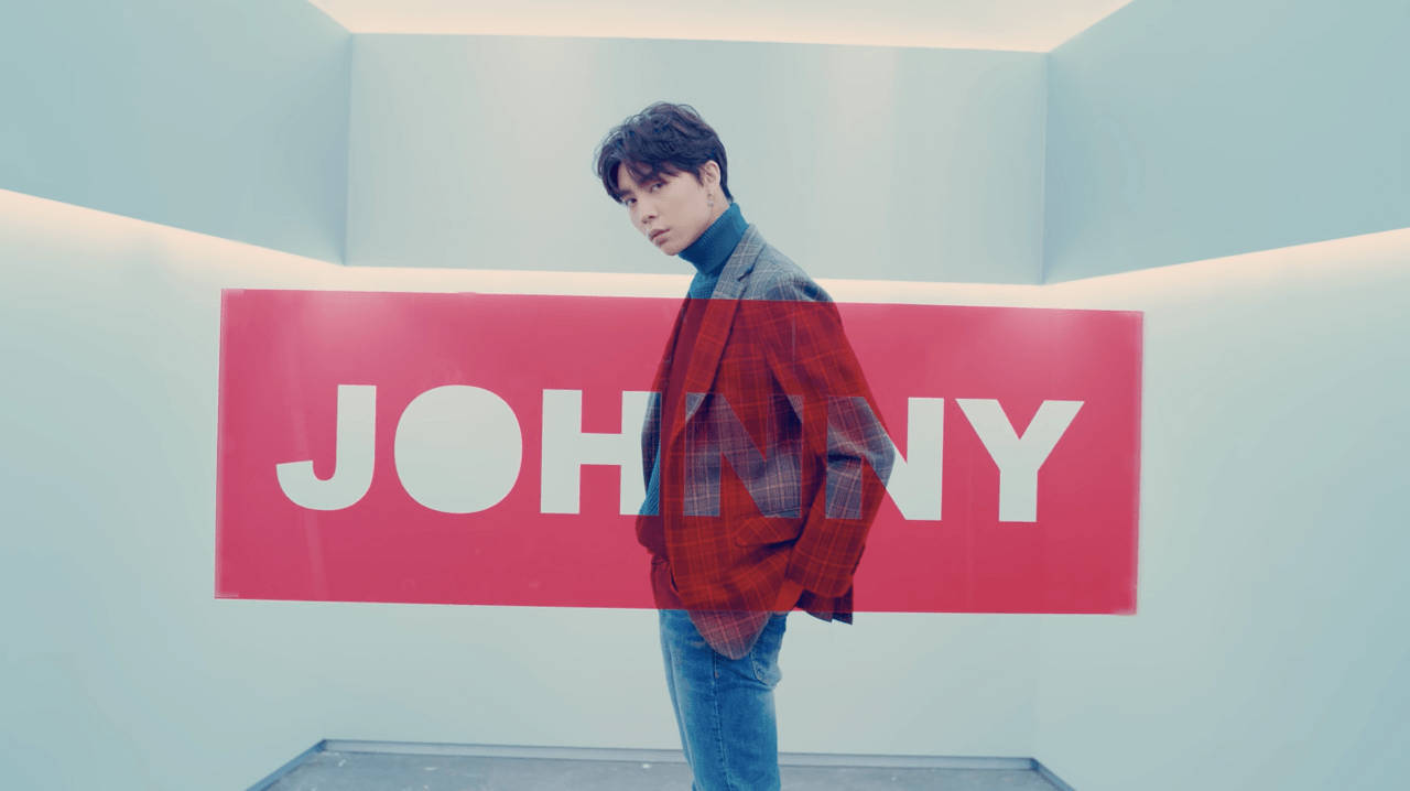 年輕男子身穿紅色格紋外套在白色房間內的粉紅色 Johnny 標誌前擺姿勢。