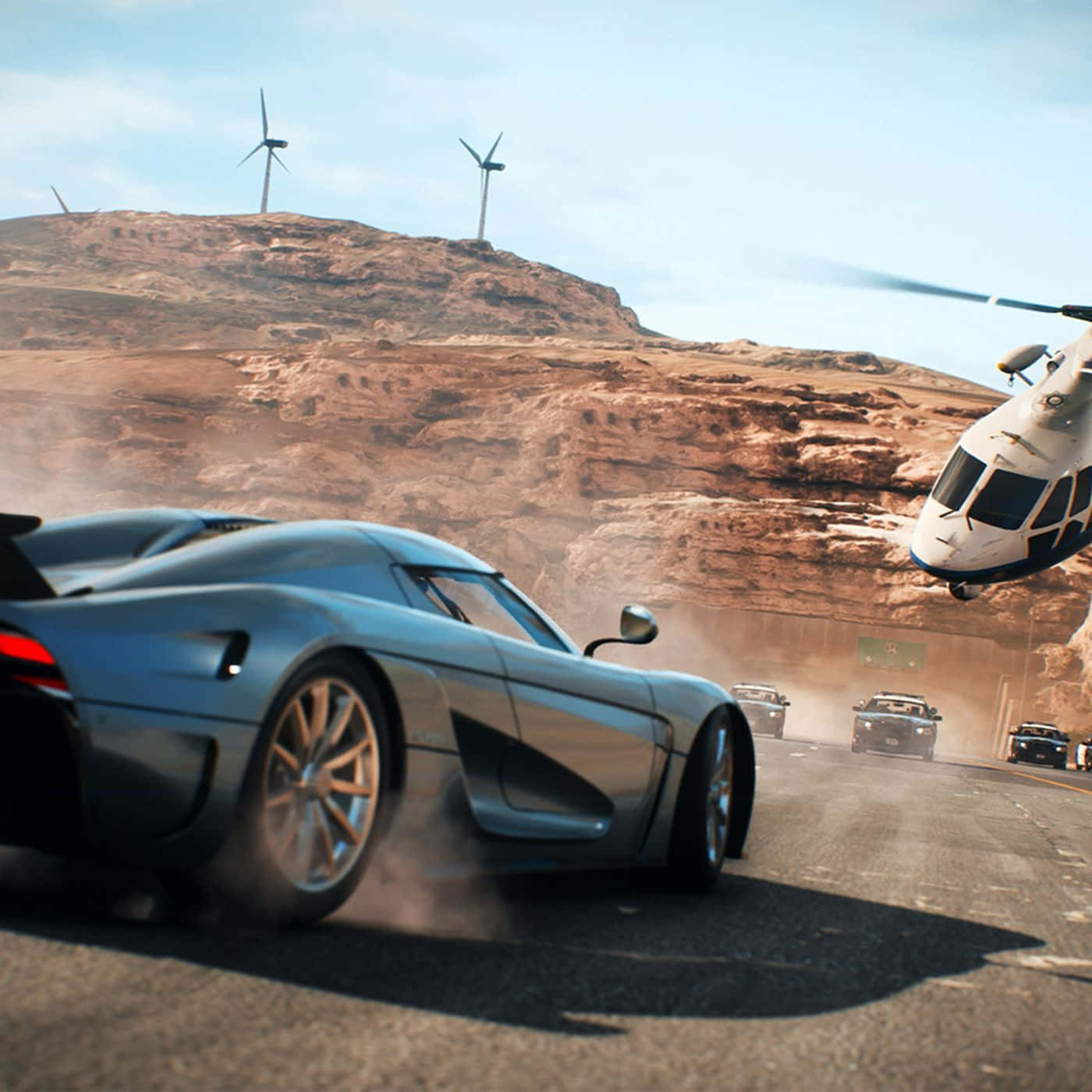 《Need for Speed Payback》中的心跳加速動作 壁紙 — 一架時尚的銀色跑車在沙漠公路上疾馳，附近有直升機盤旋，遠處山丘上有風力發電機。