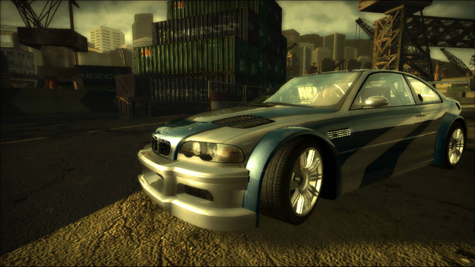 《Need For Speed》灰色BMW M3 GTR停泊 壁紙 — 一架定製的銀色和藍色BMW M3跑車停泊在一個佈滿貨櫃和起重機的粗糙城市碼頭，背景是朦朧的日落天空。