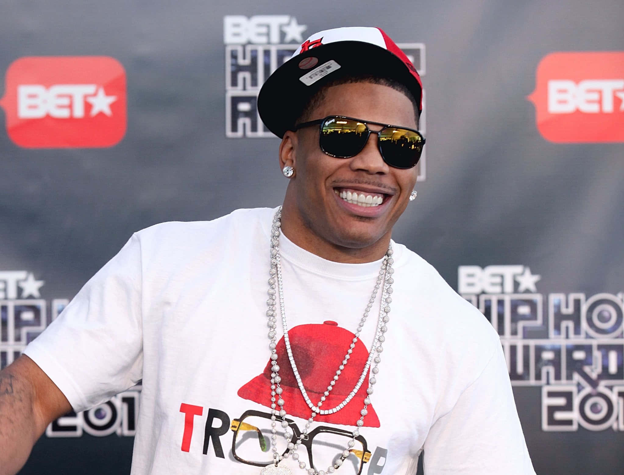 Nelly 在 2011 年 BET 饒舌音樂獎上微笑 壁紙 — 一位微笑的男子戴著紅白帽子和太陽眼鏡，在BET Hip Hop Awards上穿著圖案T恤和分層珍珠項鍊擺姿勢。