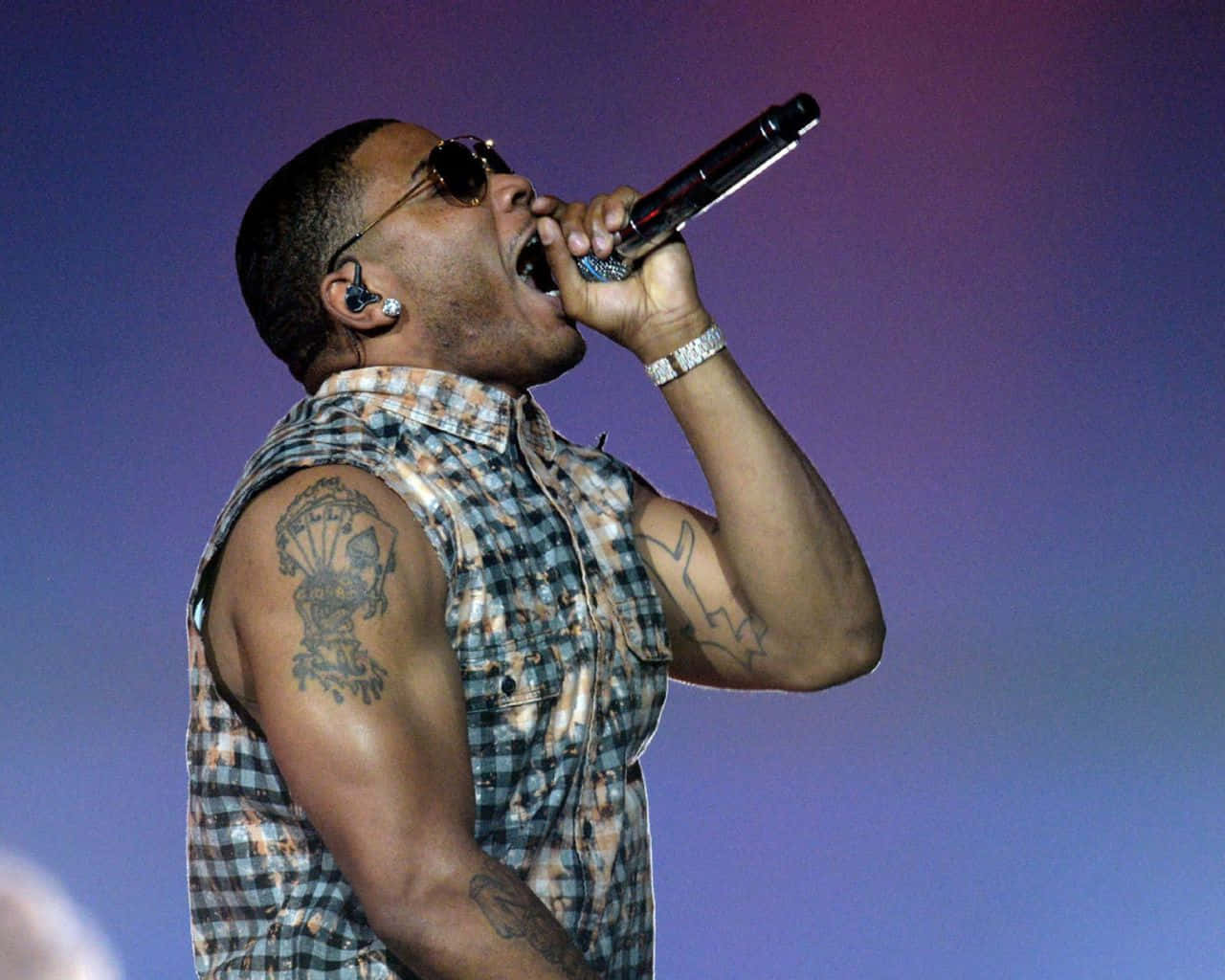 Nelly 持麥克風演唱於演唱會 壁紙 — 一位有紋身和太陽眼鏡的男歌手在紫色舞台燈光下熱情地對著麥克風表演。