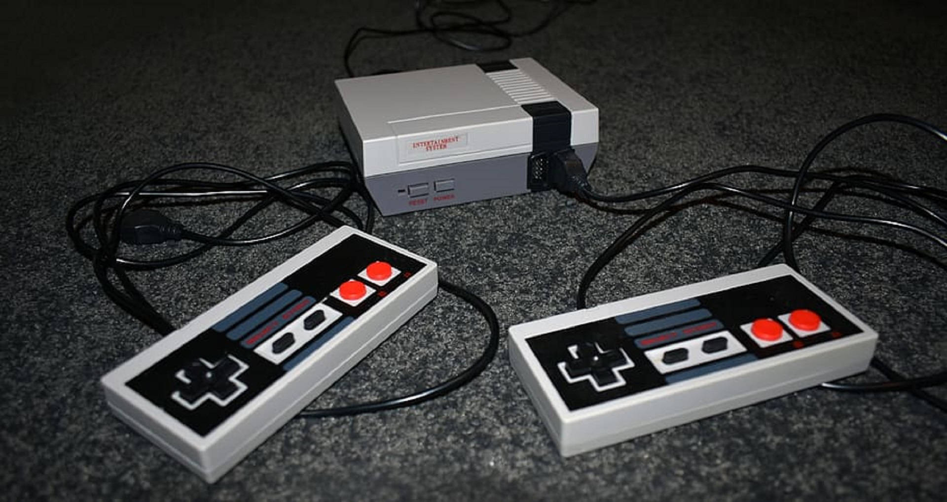 NES置於灰色地毯上 壁紙 — 復古灰色 Nintendo Entertainment System 主機連同兩個控制器放在深色地毯上。