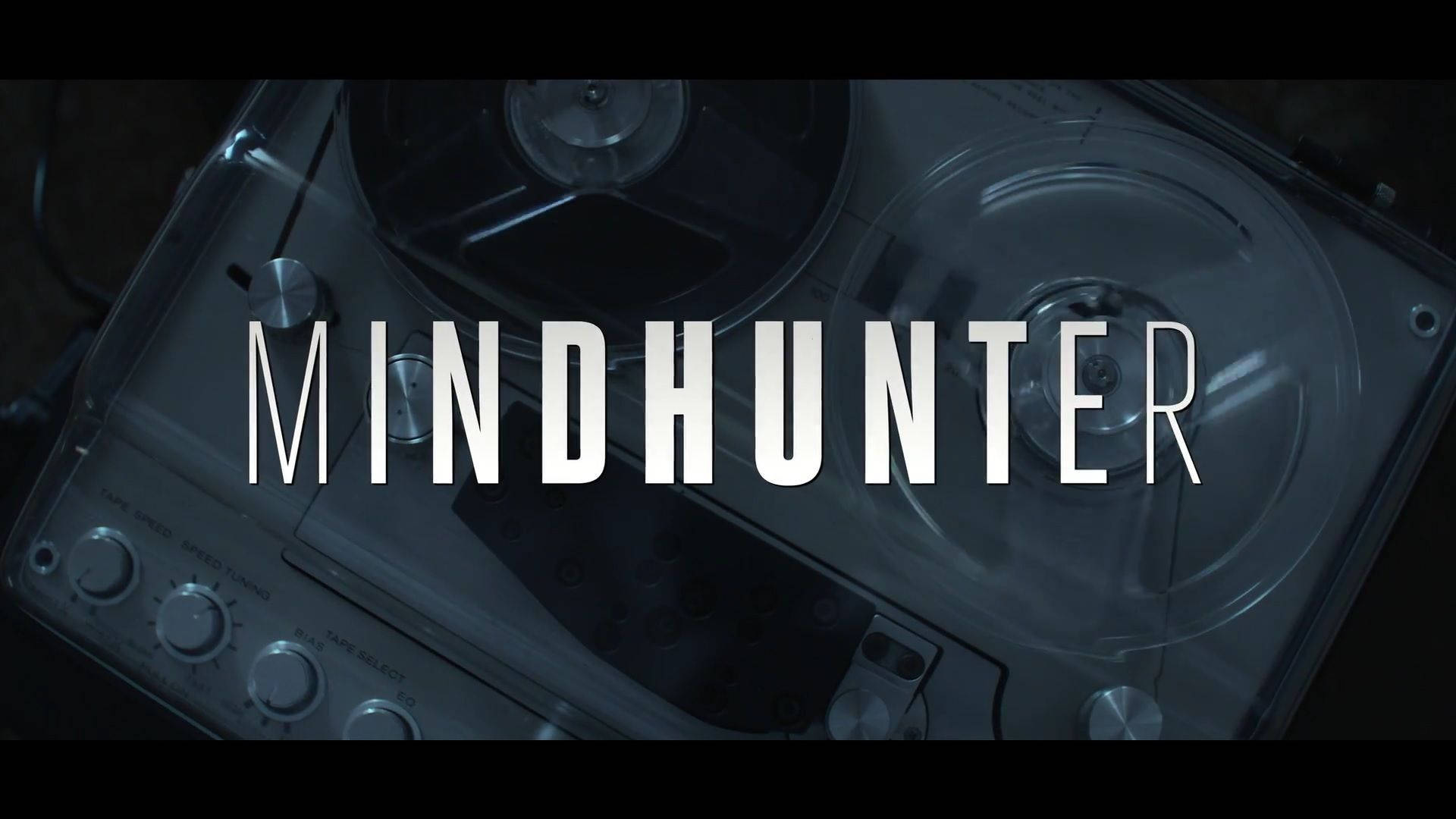 Netflix《Mindhunter》官方開場字幕 壁紙 — 一部復古的開盤式錄音機在昏暗燈光下發光，上面覆蓋著粗體白色字體的標題「MINDHUNTER」。