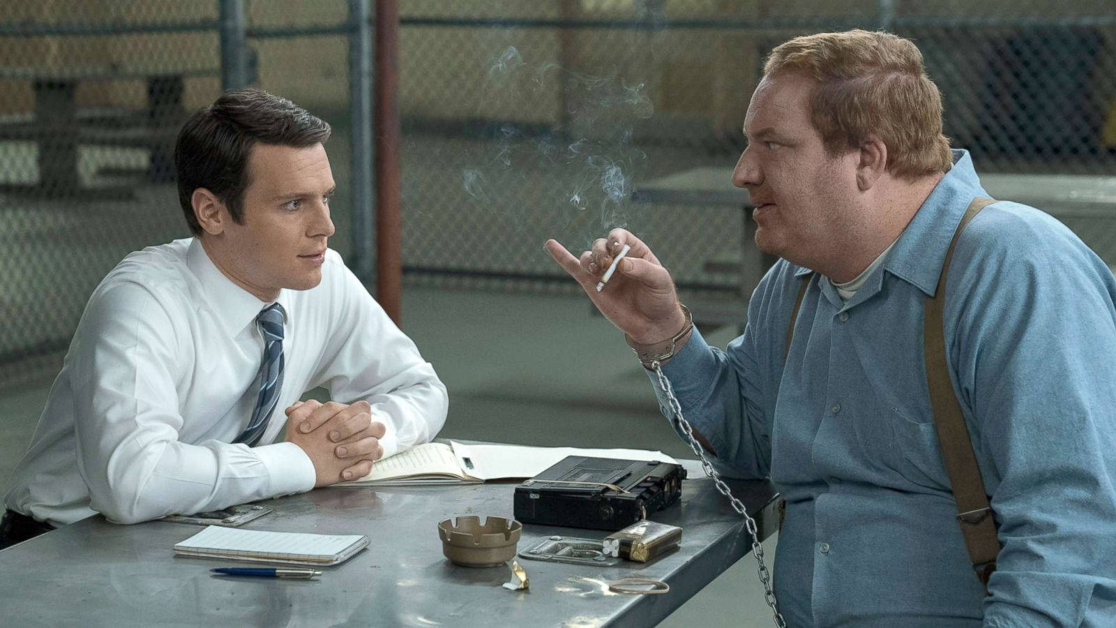 Netflix《Mindhunter》傑瑞·布魯多斯 壁紙 — 兩名男子在監獄庭院中隔著金屬桌對坐，一人抽煙，另一人專心聆聽。
