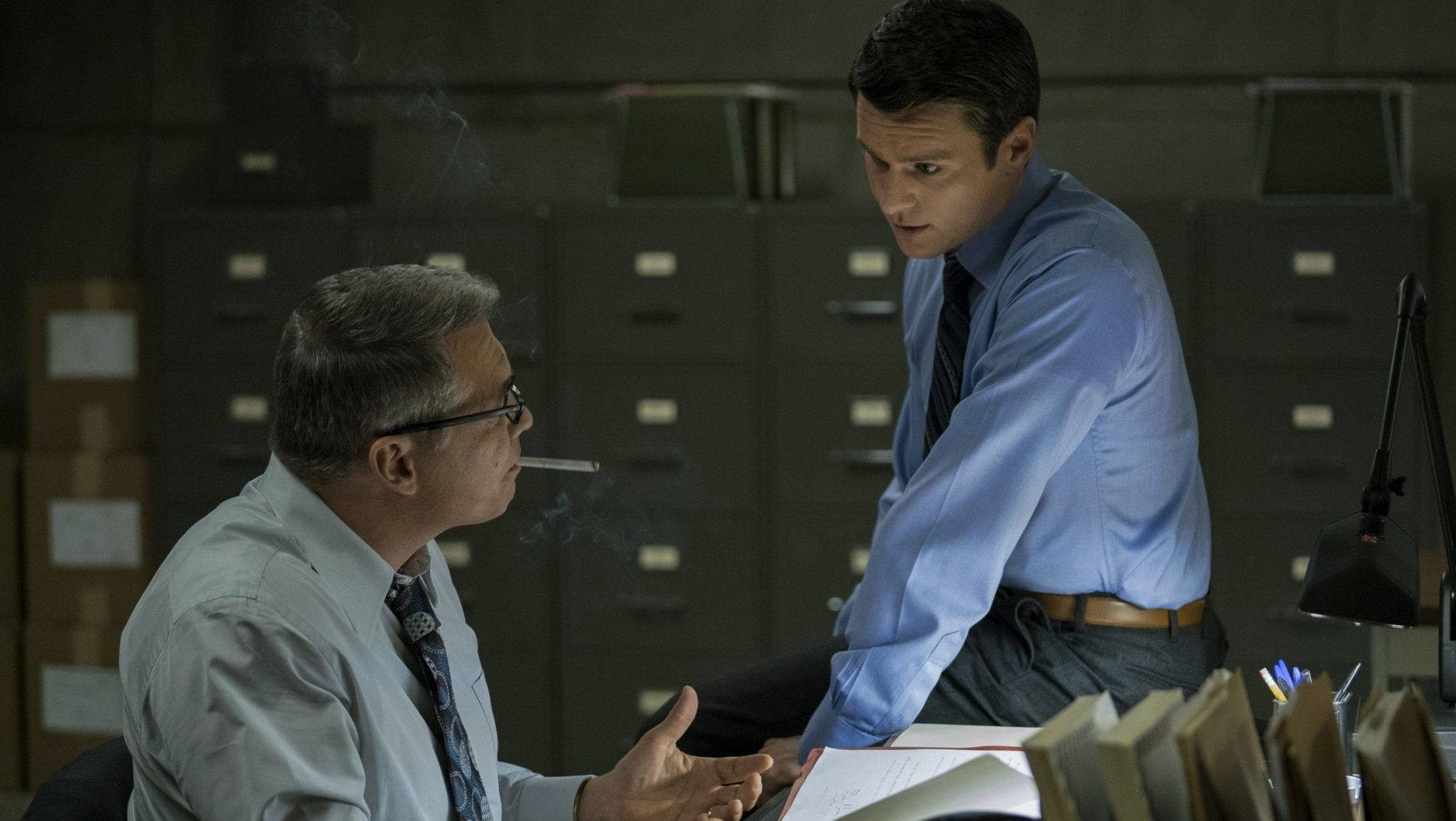 Netflix《Mindhunter》劇中比爾·坦奇抽菸場景 壁紙 — 兩個男人在昏暗的辦公室裡，一個抽煙並俯身靠在桌子上，另一個專心聆聽，周圍是文件櫃。