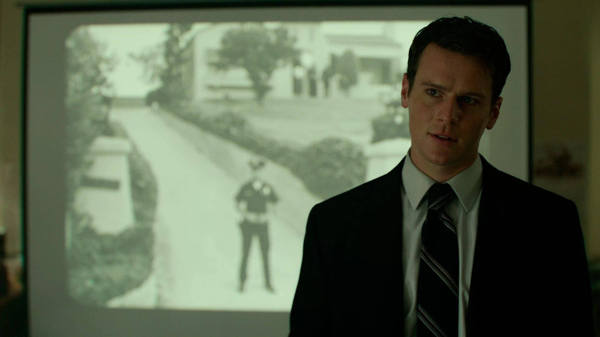 Netflix《Mindhunter》FBI探員霍爾登·福特 壁紙 — 一名身穿西裝的男子專注地凝視著投射出來的黑白保安錄像，畫面中一名警察站在住宅街道上。